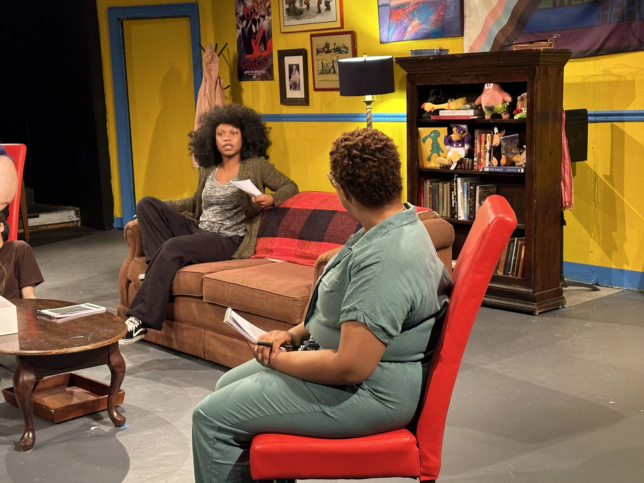 Two women have a conversation on stage with a colorful, artistic backdrop and bookshelf behind them. One woman with voluminous curly hair is seated on a couch, and the other woman with short hair and glasses is seated on a red armchair.