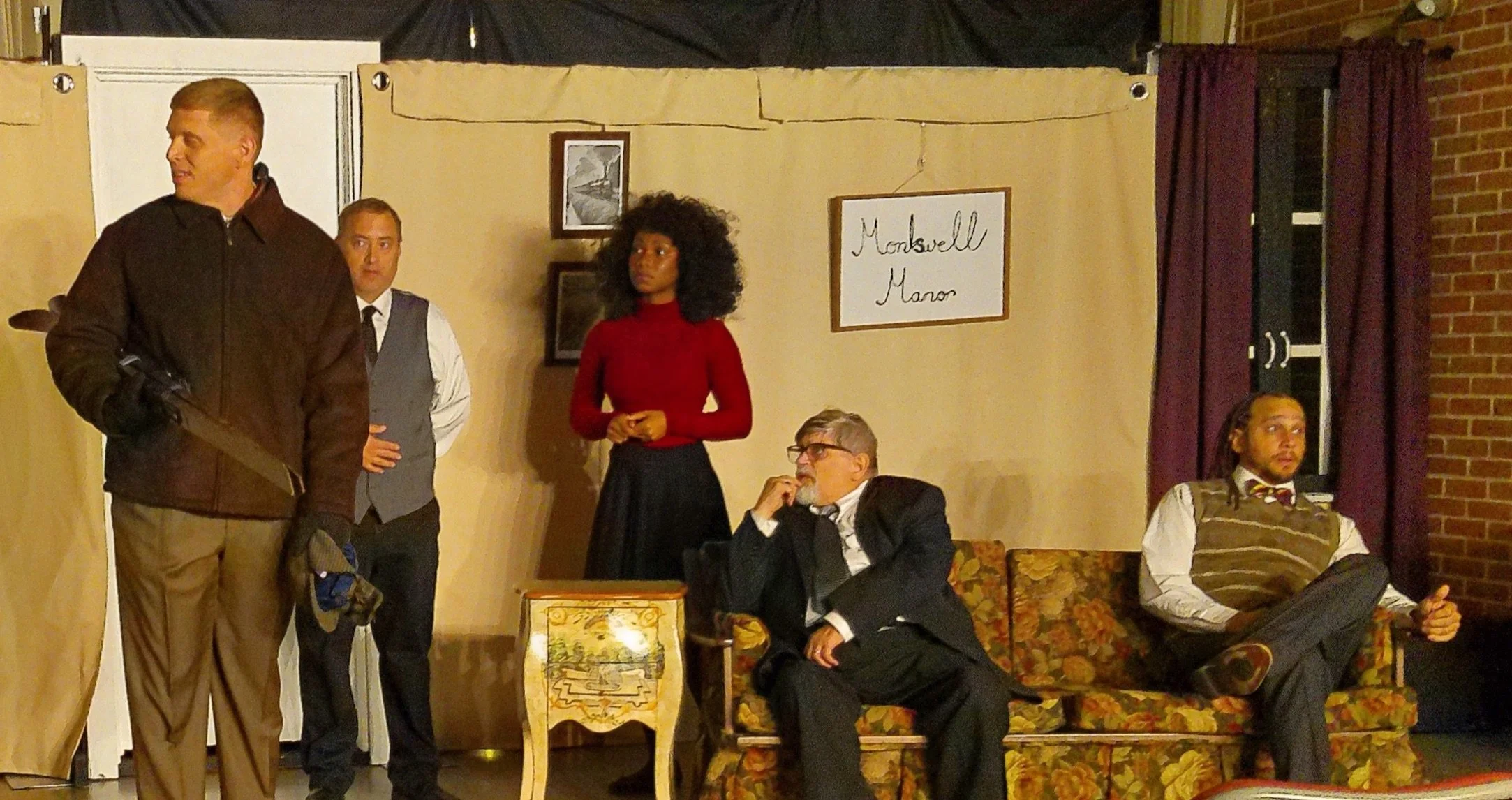 Five people in a room with a beige backdrop and purple curtains. One man is standing holding a gun, another man is sitting on a floral couch with one hand on his chin, a woman in a red turtleneck and black skirt is standing behind the couch, a man in