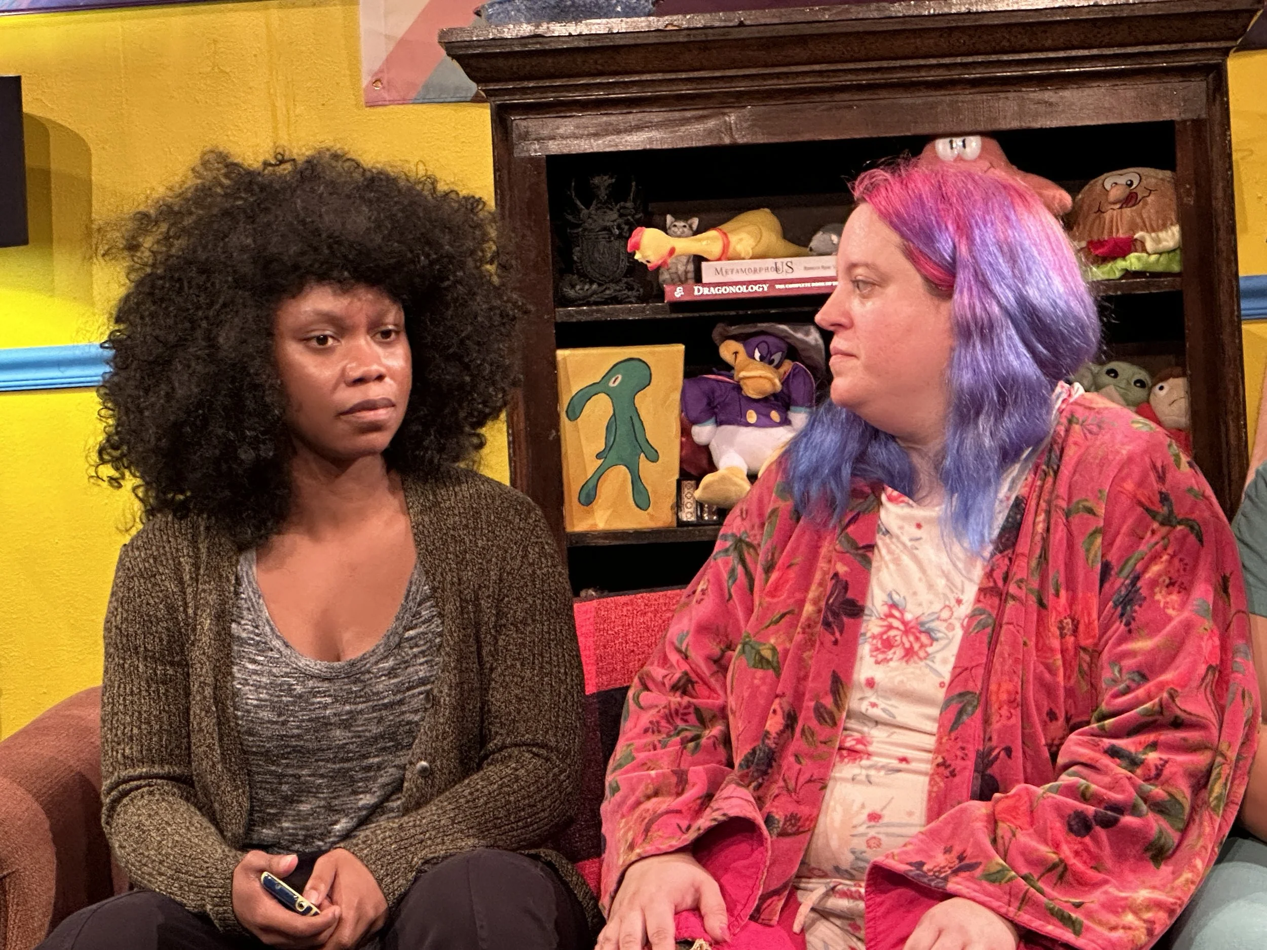 Two women sitting and engaging in conversation in a colorful room with yellow walls and shelves filled with plush toys, books, and art.