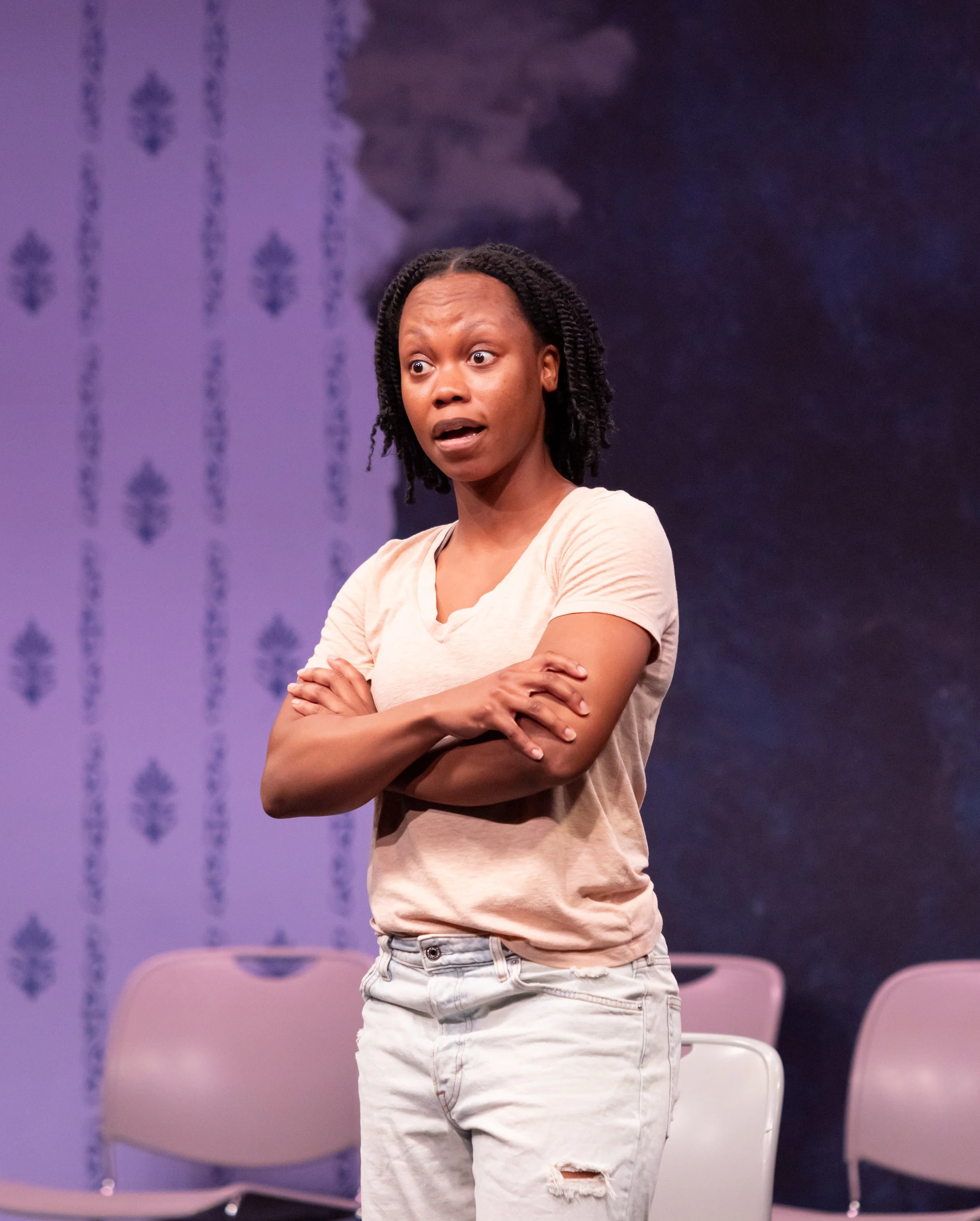 A woman with short curly hair, wearing a light beige t-shirt and ripped white jeans, standing with arms crossed, looking surprised or concerned, on a stage with purple patterned wallpaper and empty chairs in the background.