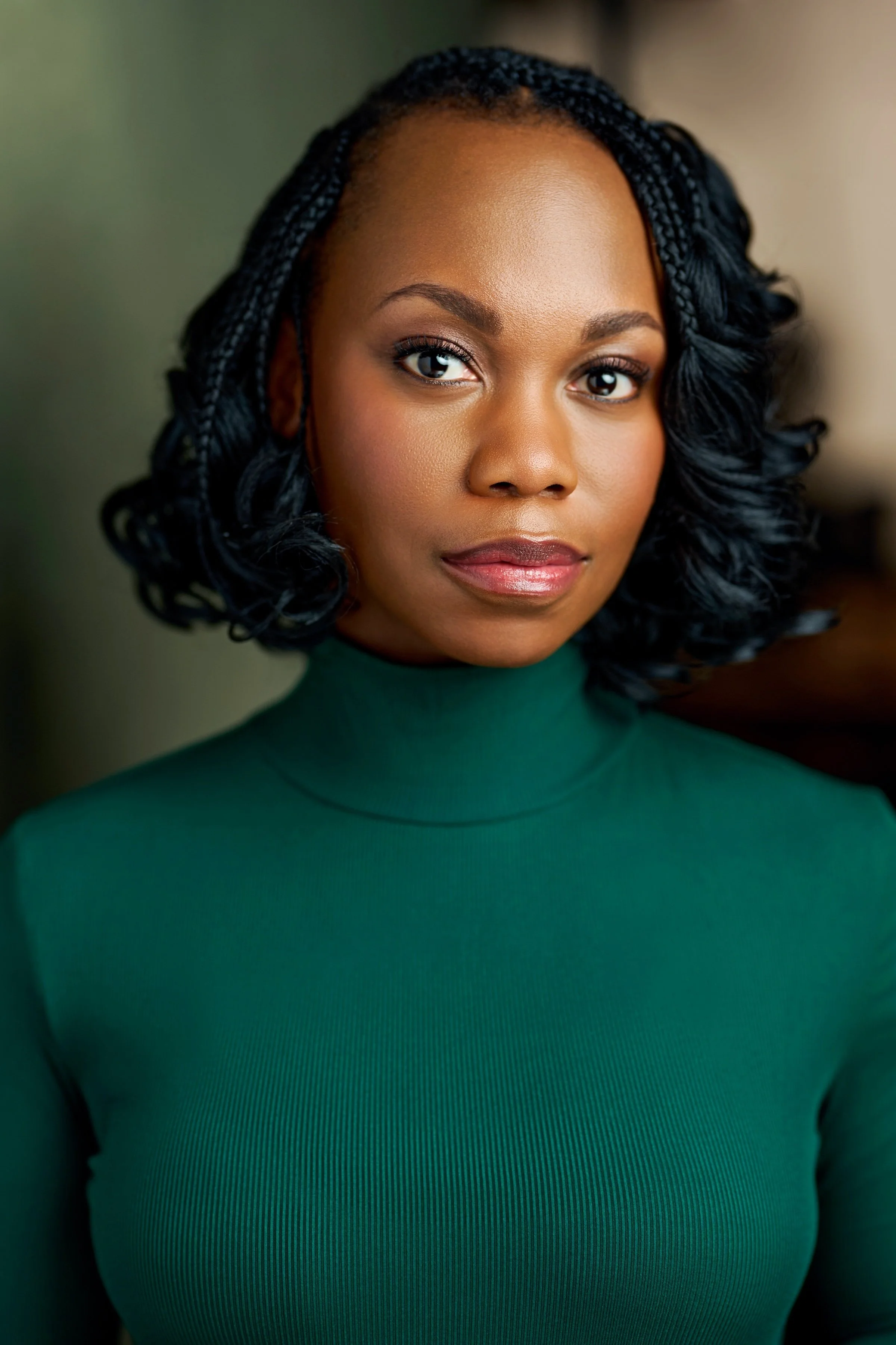 Close-up portrait of a woman with dark, curly shoulder-length hair, wearing a green turtleneck sweater, looking directly at the camera with a neutral expression.