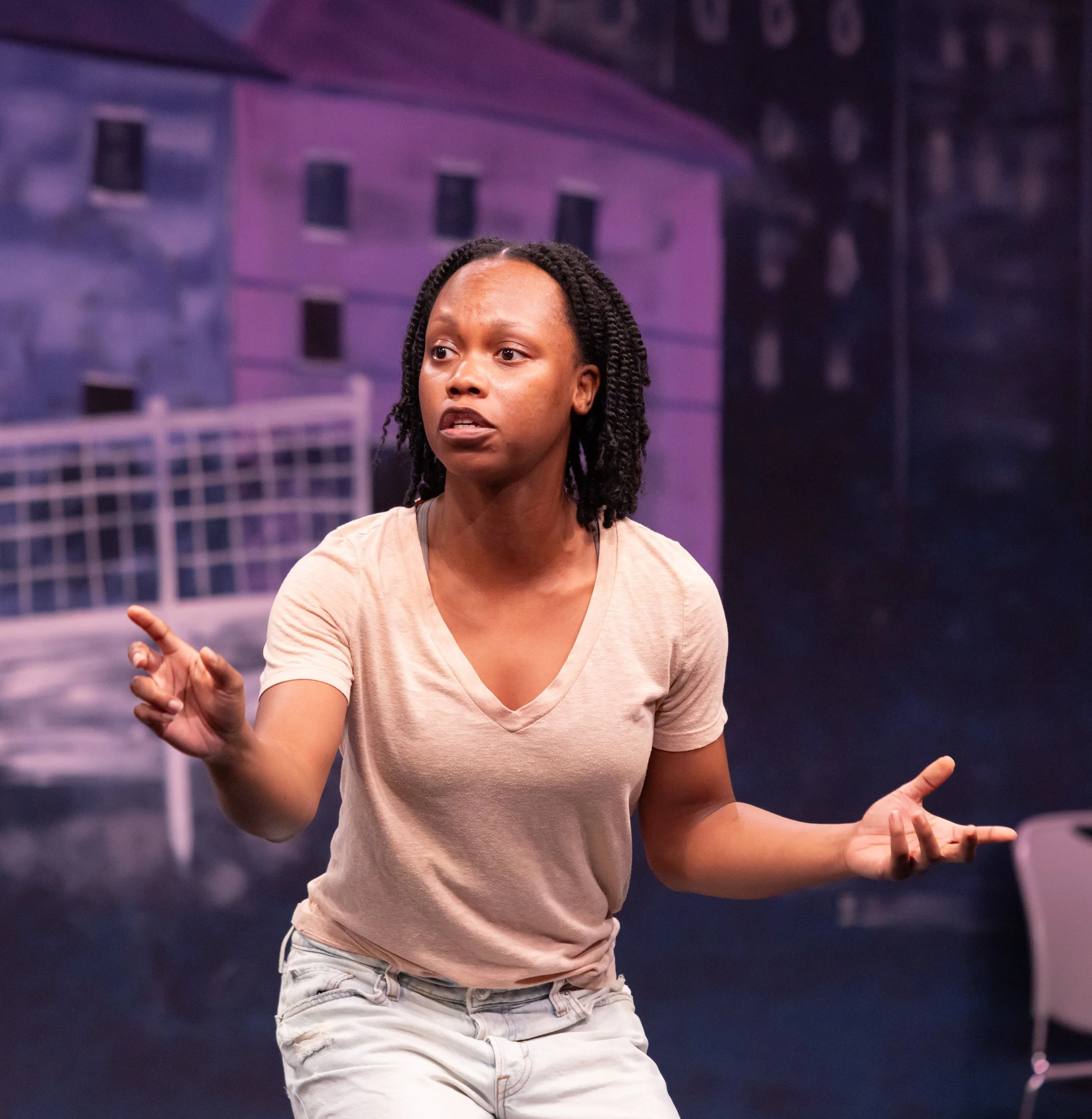 A woman with twists in a beige t-shirt and light pants is acting in a theatrical production, gesturing with her hands against a cityscape backdrop.