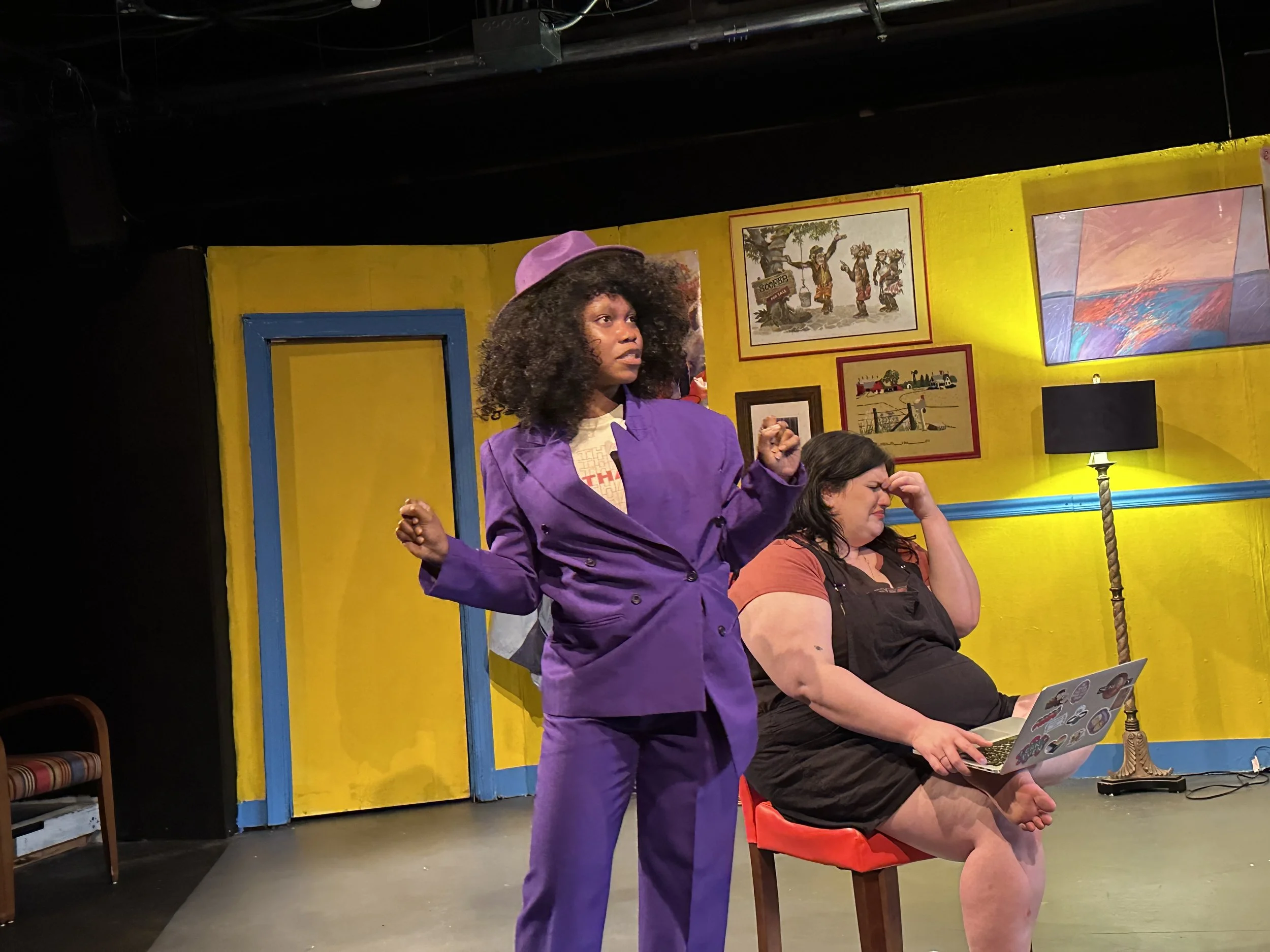 Two women on stage, one standing and the other sitting. The standing woman wears a purple suit, hat, and has curly hair. The seated woman wears a black dress, has long dark hair, and is holding a laptop. The background features colorful paintings and