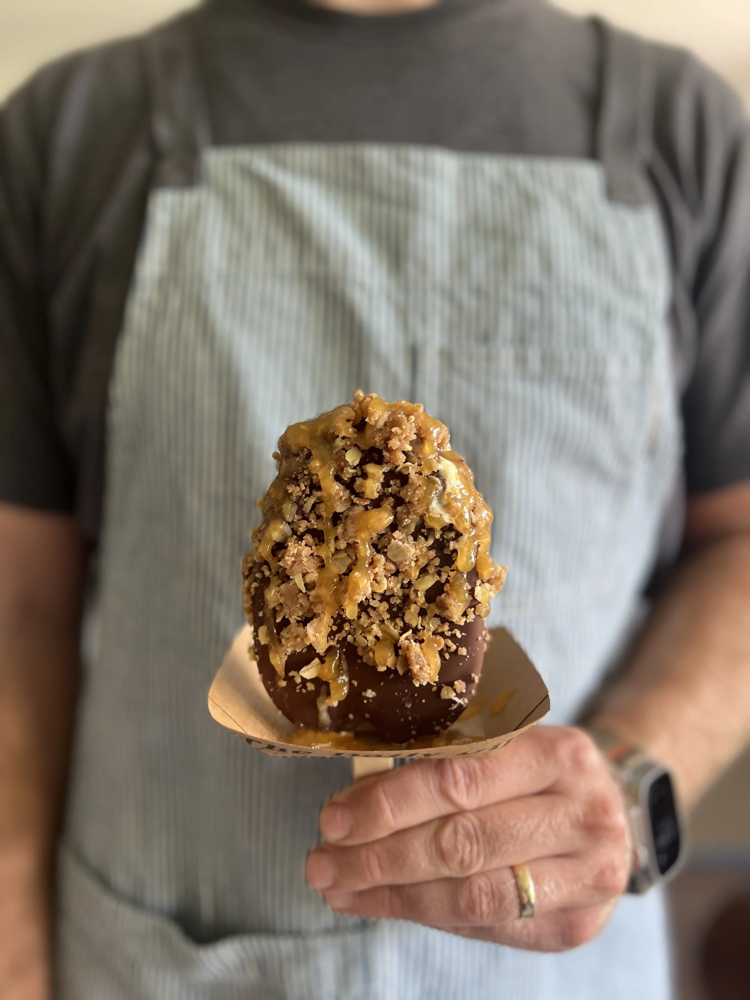 A hand holding an icecream dipped in chocolate and covered in crumble and peach drizzle