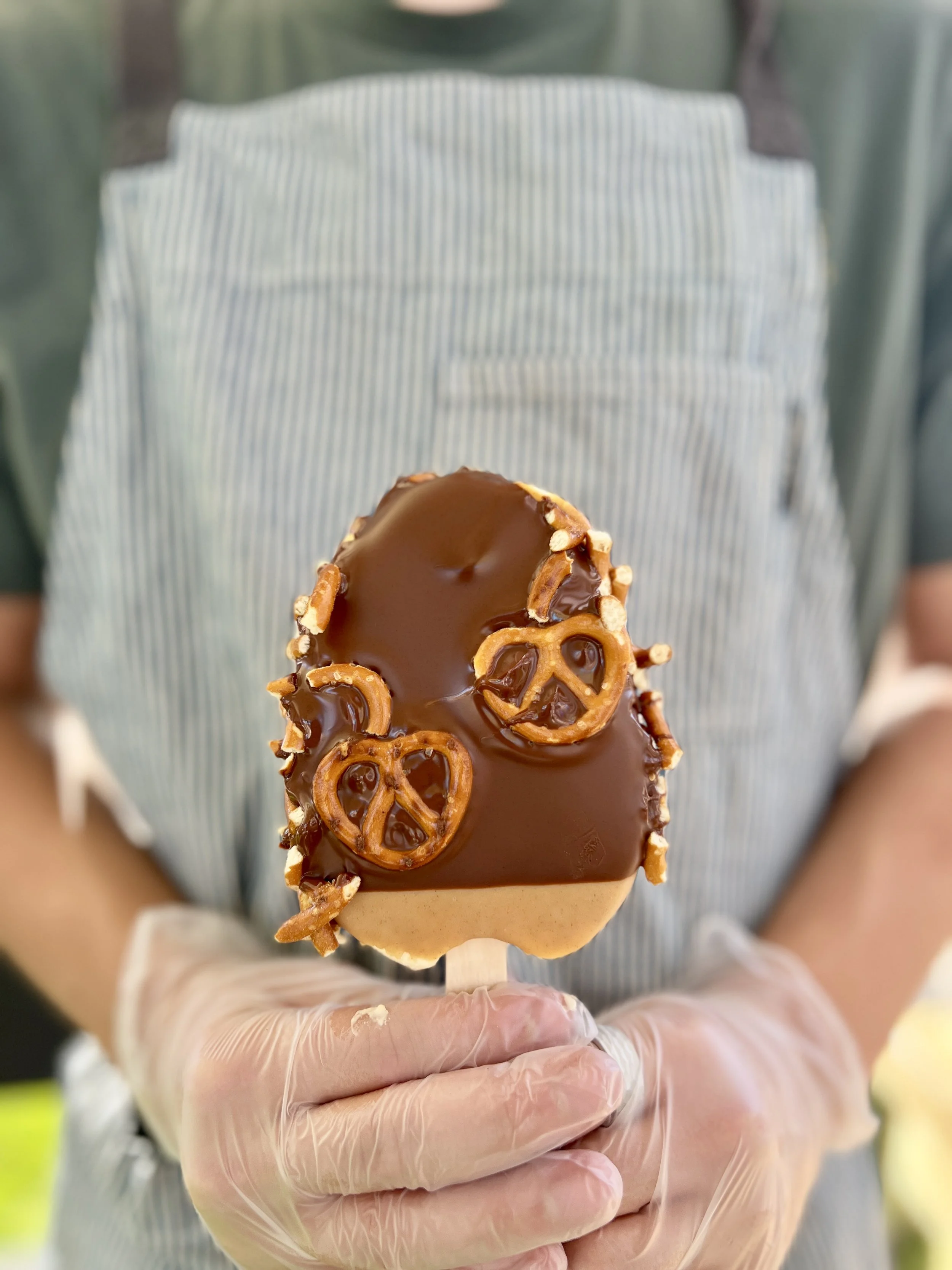Person wearing a dark shirt and striped apron holding a caramel apple covered in crushed nuts.