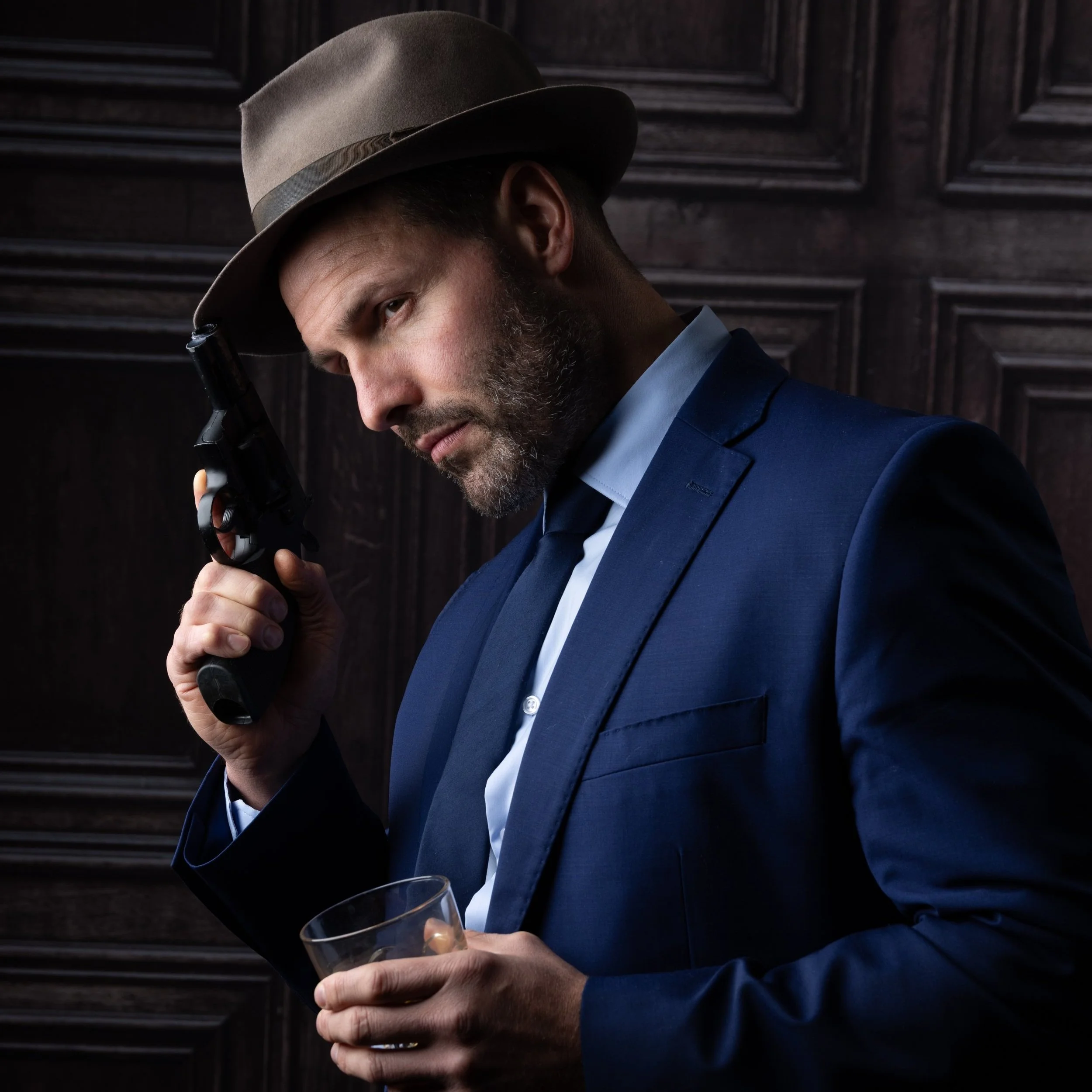 A man in a blue suit and fedora hat holding a gun to his temple, with a glass of drink in his other hand, in front of dark wood-paneled walls.