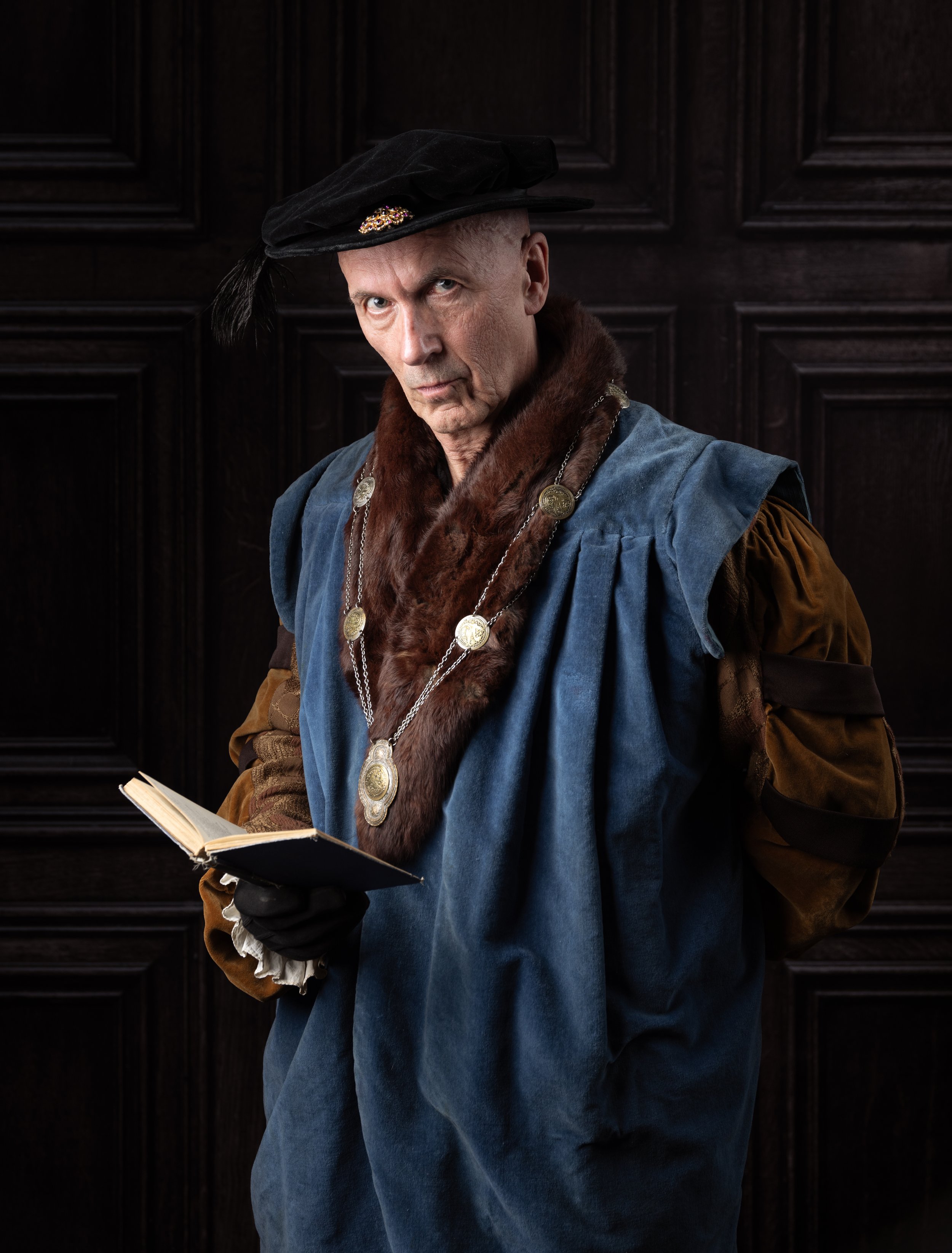 A man dressed in historical Renaissance-style clothing with a velvet hat, fur collar, and medallion necklaces, holding a small open book against a dark wooden paneled background.