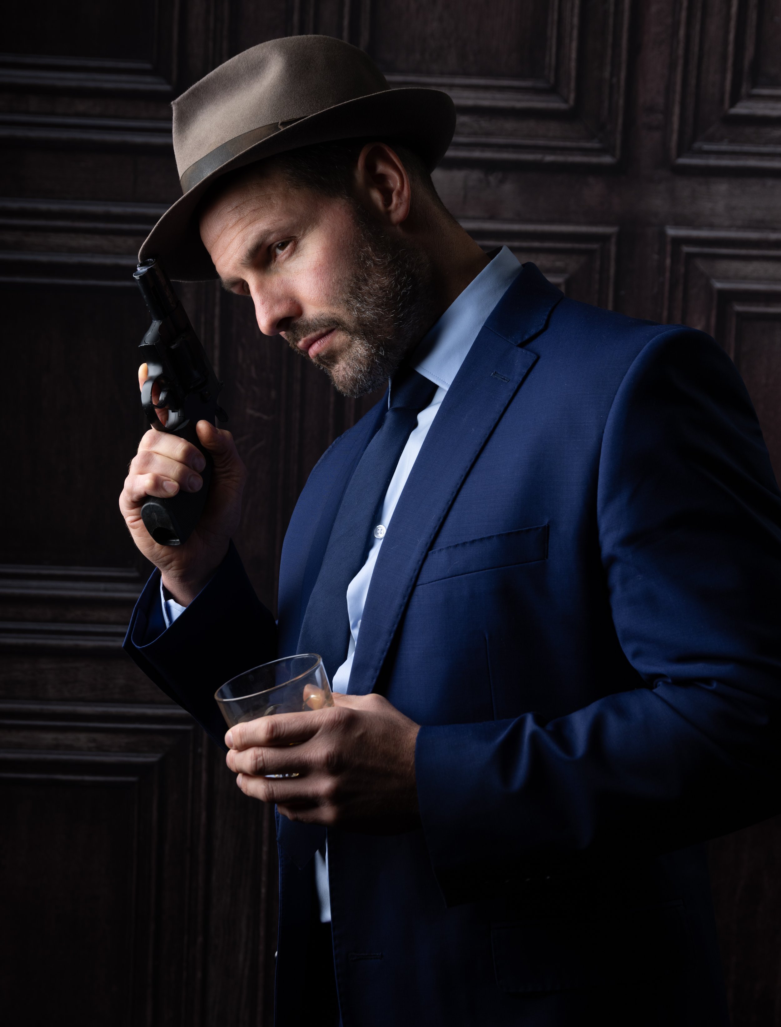 Man in a blue suit and brown fedora hat holding a pistol to his temple, with a glass of whiskey in his other hand, standing against a dark wooden background.