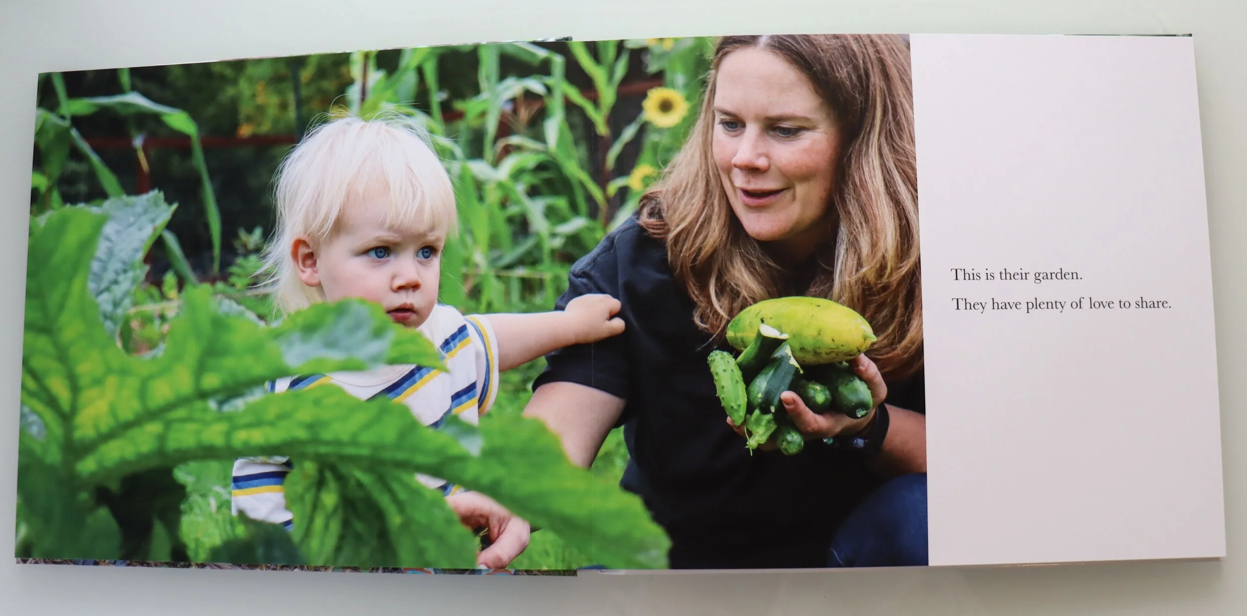A woman and a young child are in a garden, surrounded by green plants. The woman is holding a bunch of cucumbers and a zucchini, while the child, reaching out with an expression of curiosity, touches a leaf. The woman is smiling slightly, and the chi