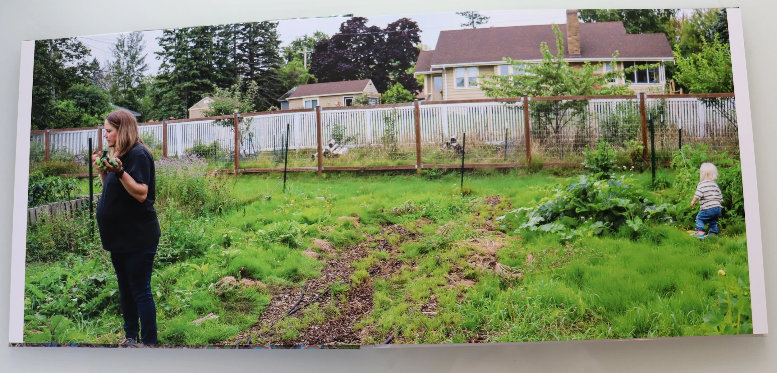 A woman standing in a garden holding a small vegetable, with a young child exploring plants nearby. The garden has a mix of green plants and soil, surrounded by a wooden fence, and houses with trees are visible in the background.