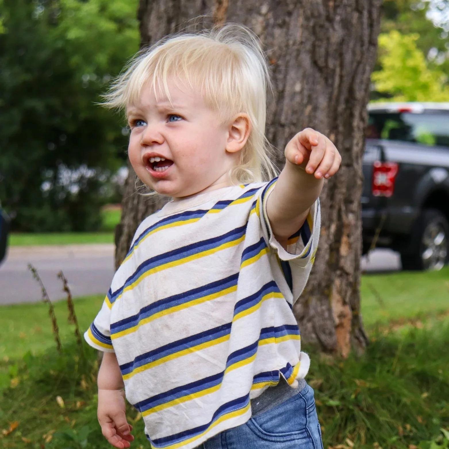 A young blonde-haired child with blue eyes wearing a yellow, blue, and white striped t-shirt and blue jeans standing outdoors next to a tree. The child has an expressive face, with mouth slightly open and one arm extended outward. Green trees and a p
