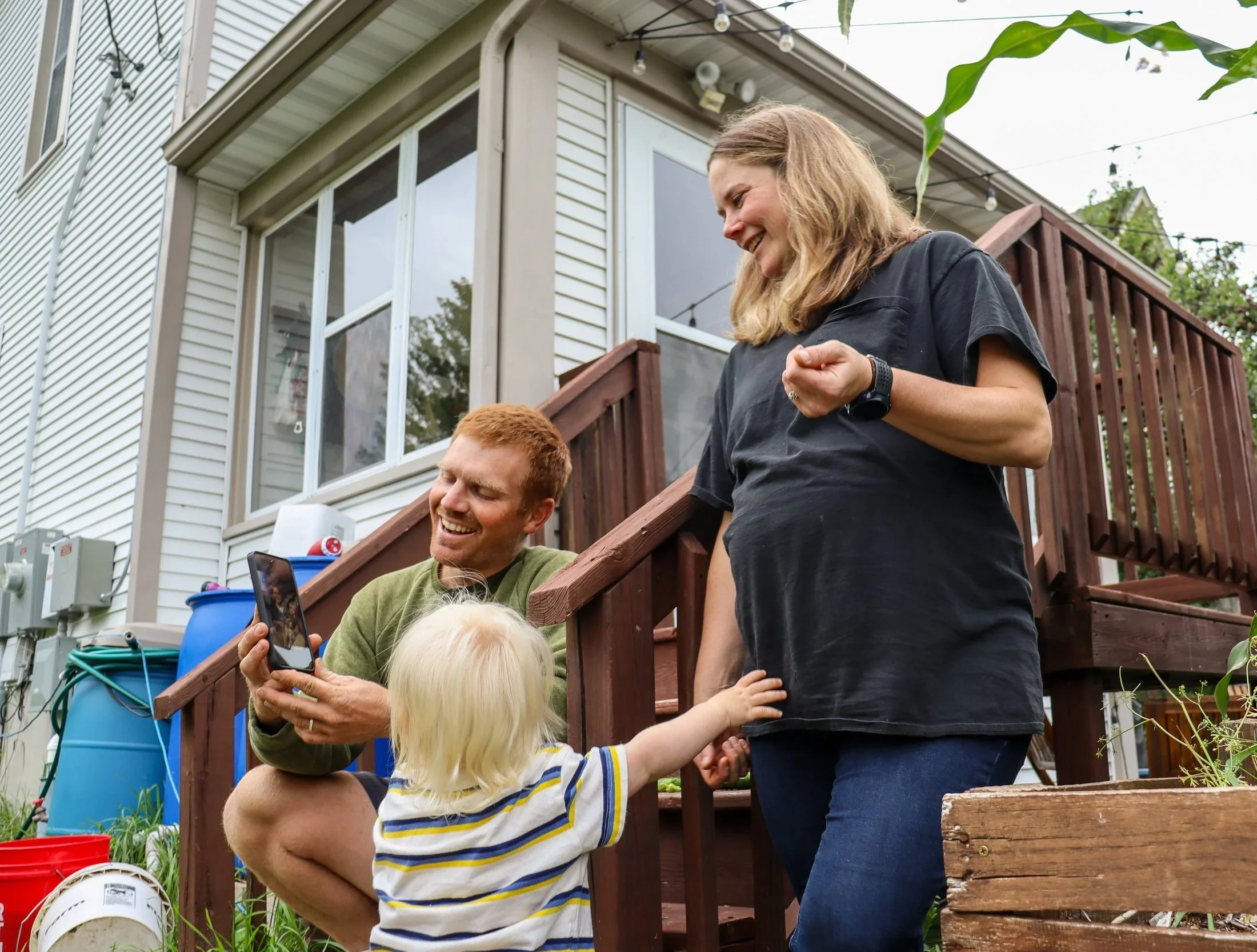 A woman and a man laugh with their toddler. The woman is smiling and reaching out to the toddler, while they are looking at the man's phone. The background shows a house with large windows, a deck, and some trees, with string lights hanging overhead.