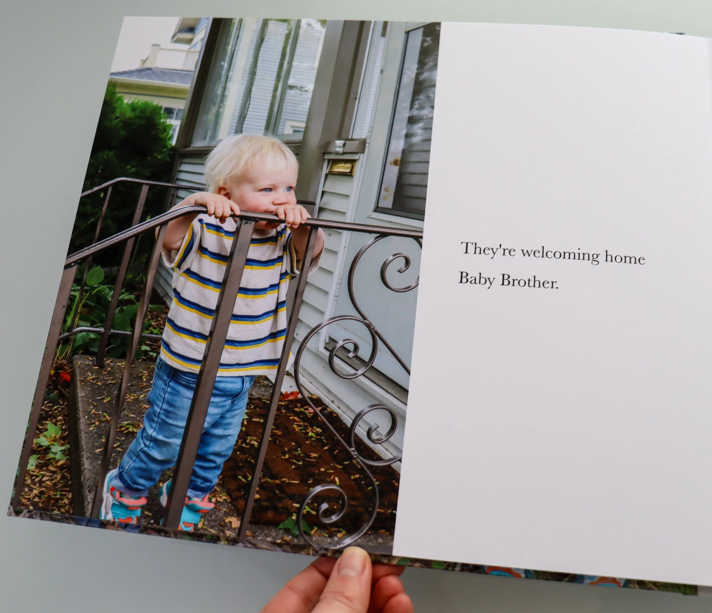 A young boy with blonde hair wearing a striped shirt is leaning on a metal railing outside a house with gray siding and multiple windows. There are green bushes and fallen leaves on the ground.