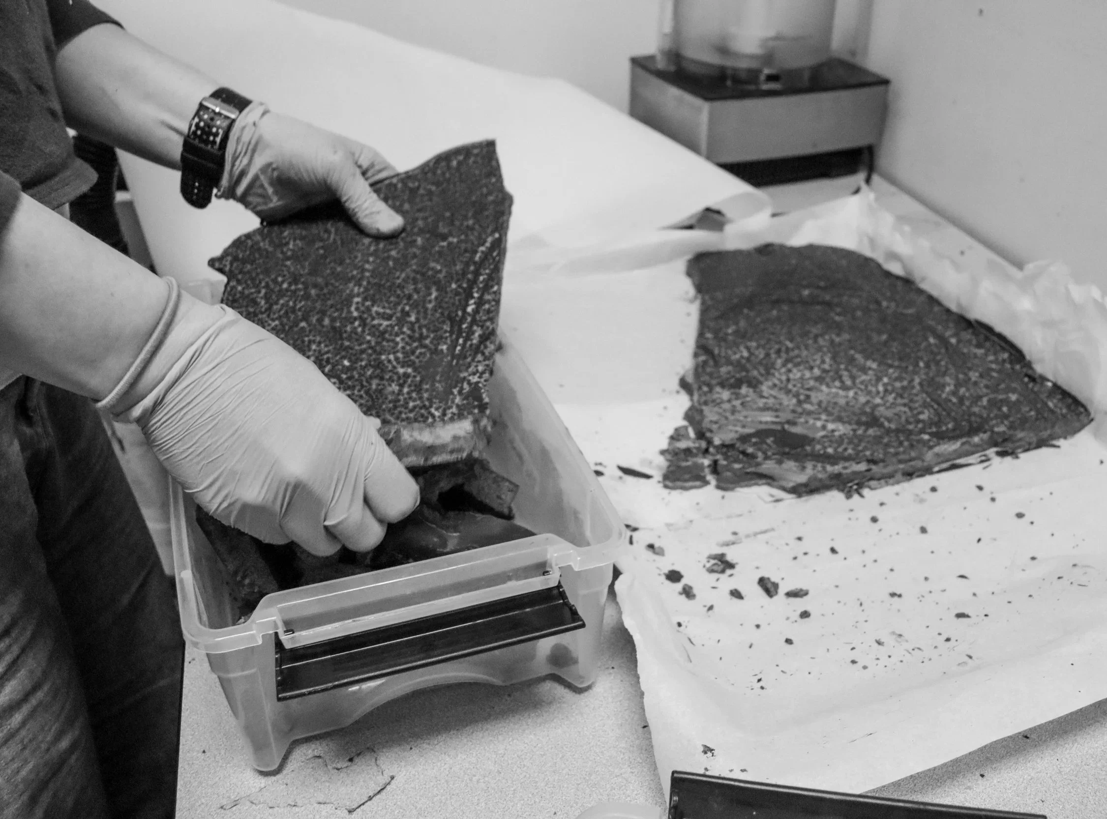 Person wearing gloves handling a large slab of chocolate, with a food processor in the background, in a monochrome setting.