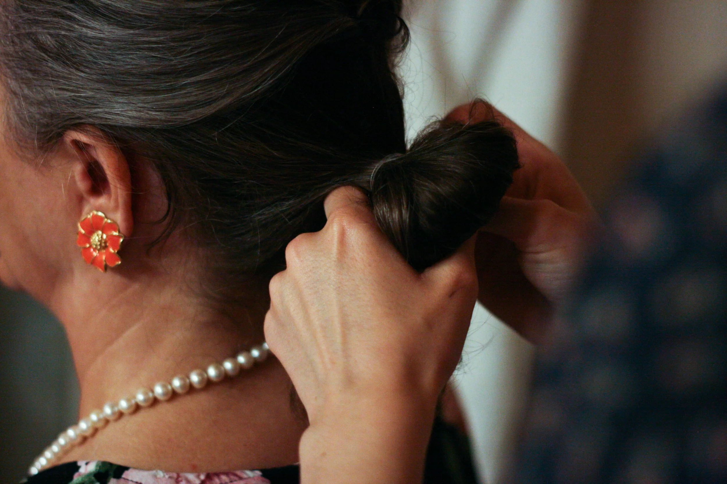 Close-up of a woman with dark hair wearing floral earrings and a pearl necklace, covering her face with her hand.