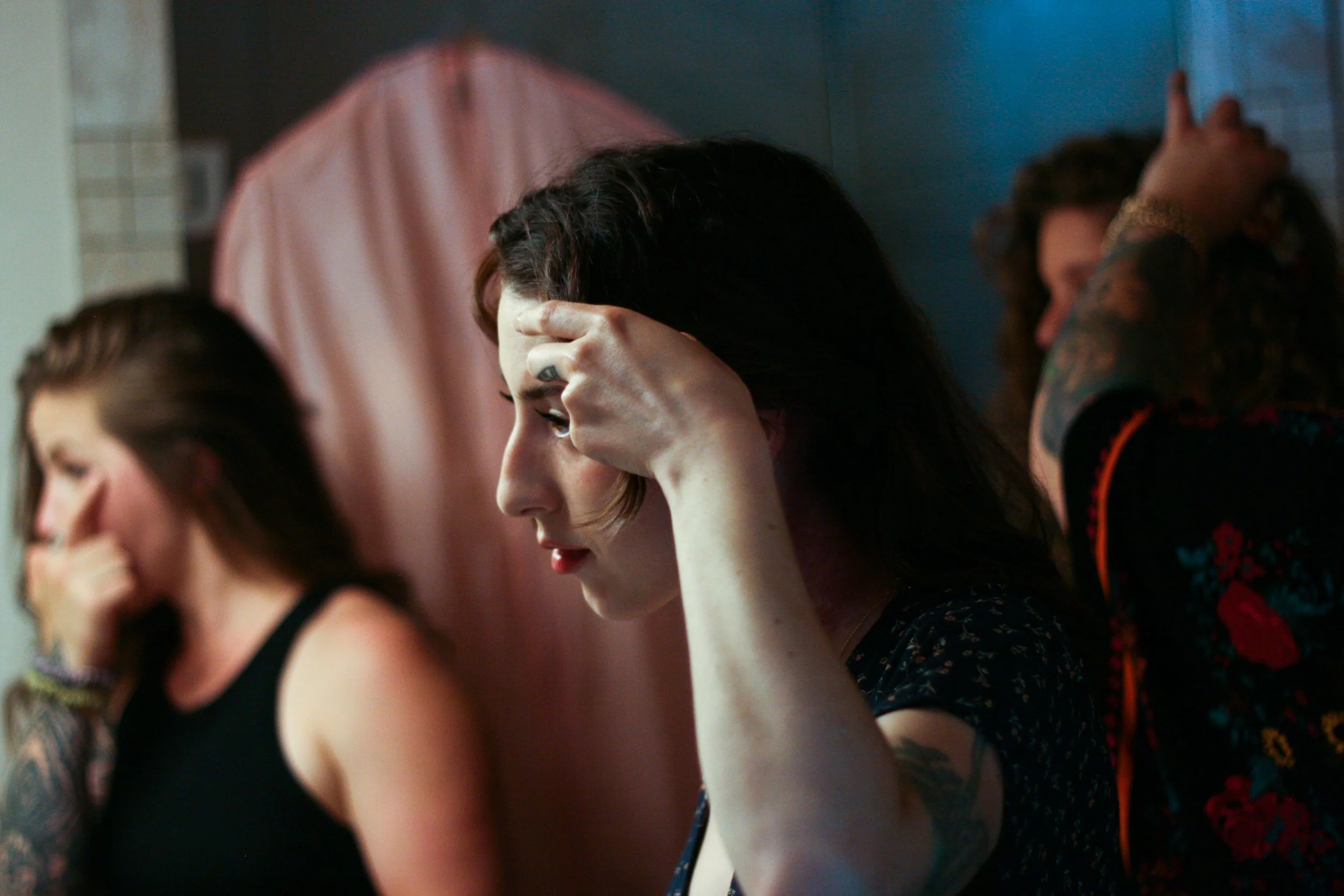 A group of women sitting at a table, one woman with dark curly hair and tattoos on her arm, holding her head with her hand in concentration.