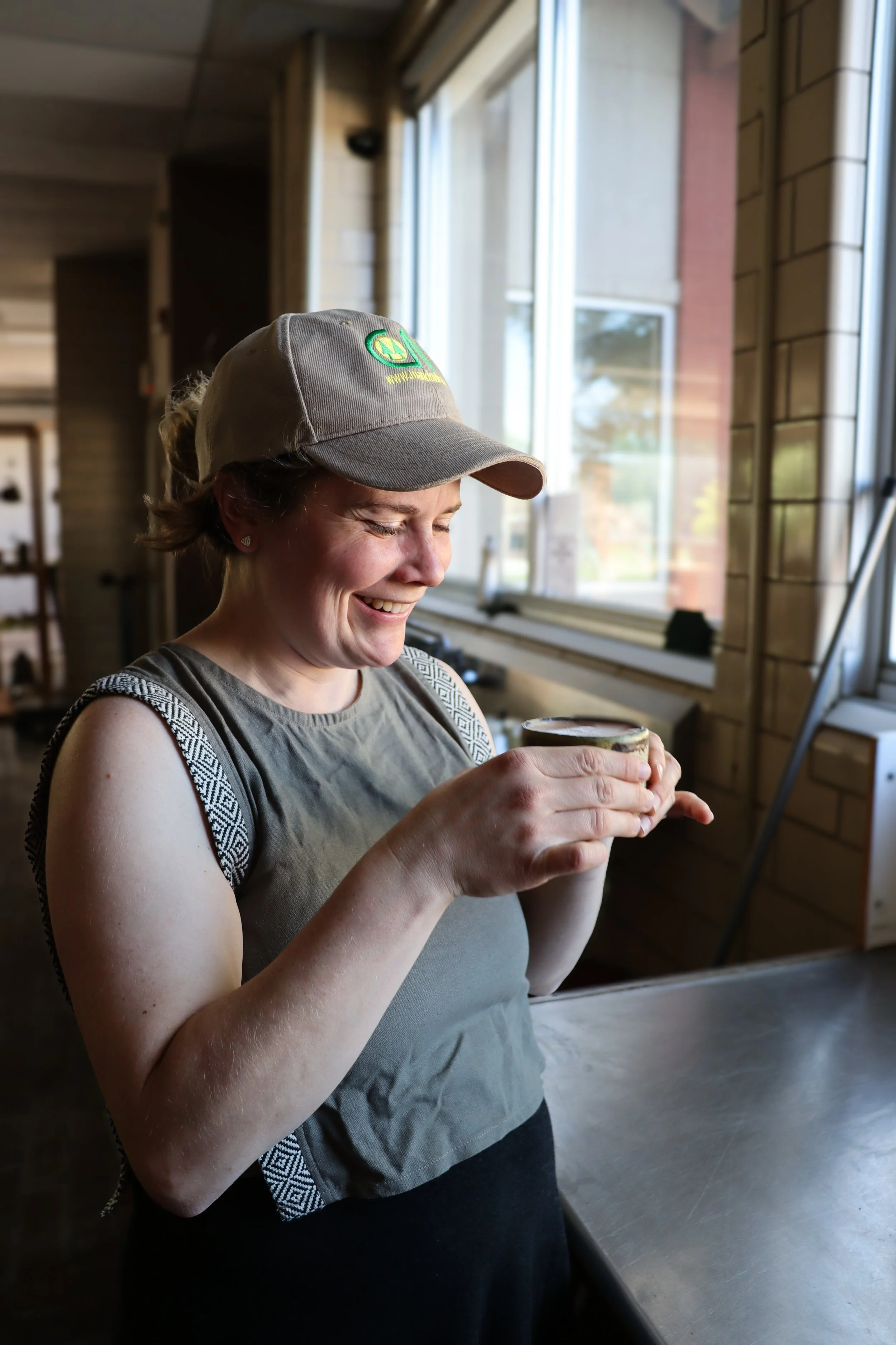 A woman wearing a tan baseball cap with a logo and a gray sleeveless top, smiling while holding a cupcake or small container, standing inside a cafe or bakery near a large window with natural light.