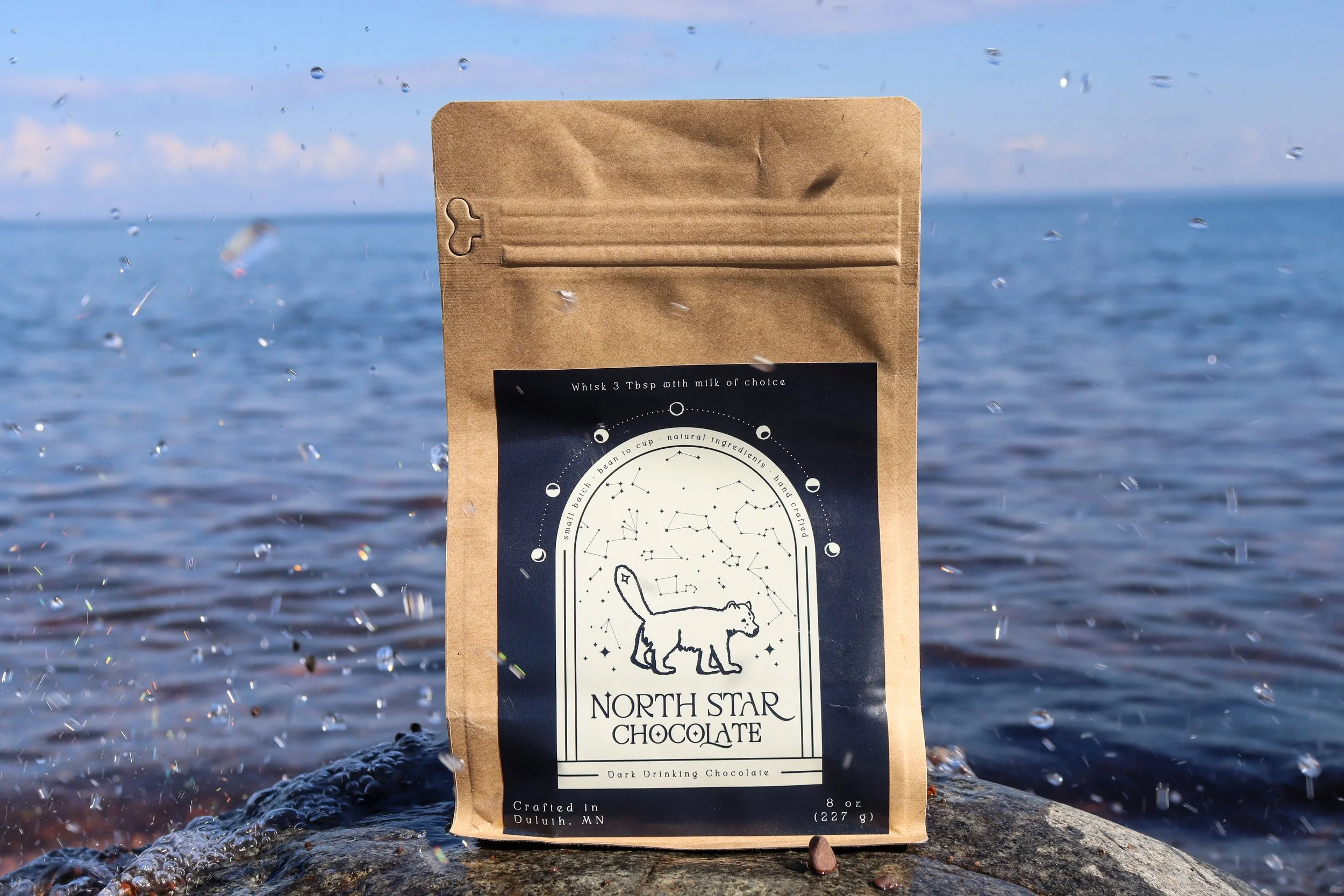 A brown paper bag of North Star Chocolate dark drinking chocolate standing on rocks at the edge of the water, with a blue sky and ocean in the background, and water droplets around the bag.