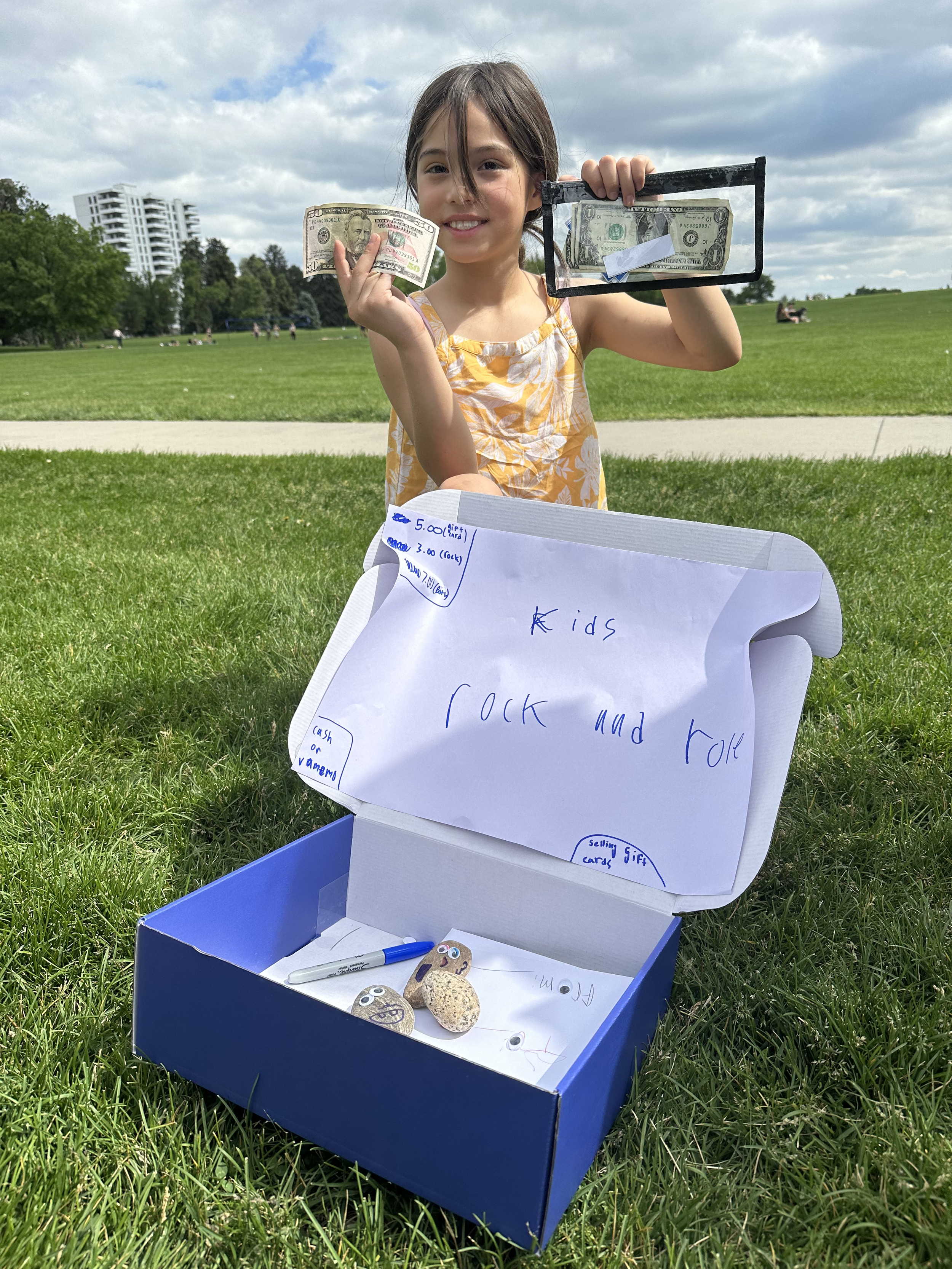 A young girl selling rocks decorated with googly eyes, holding a $10 bill and a glass case with a dollar bill, next to a sign that reads "Kids Rock and Roll" at a park on a cloudy day.