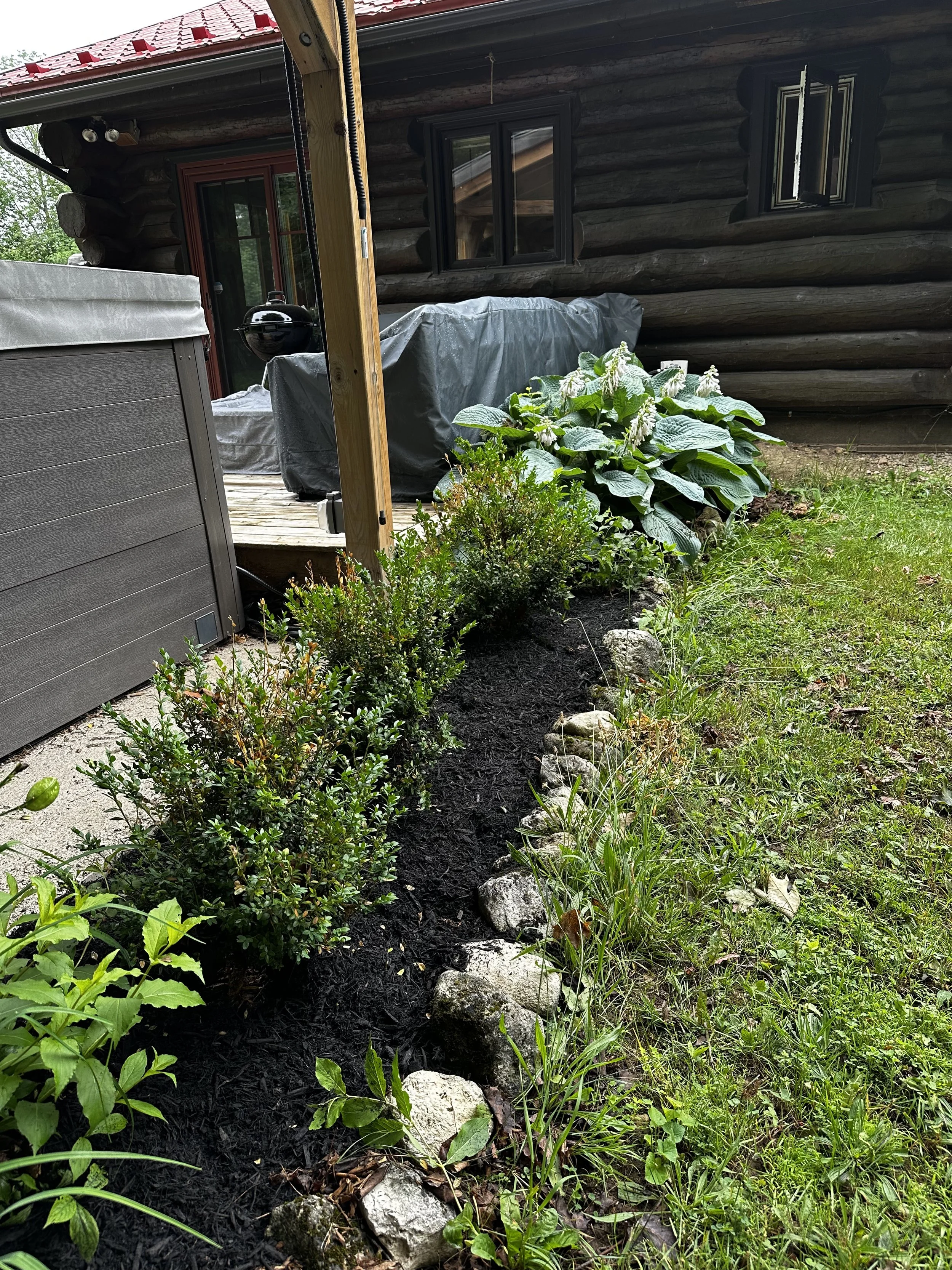 A garden bed with shrubs, flowering plants, and a row of rocks along the edge, adjacent to a log cabin house with a sliding glass door, windows, and a hot tub covered with a gray cover.