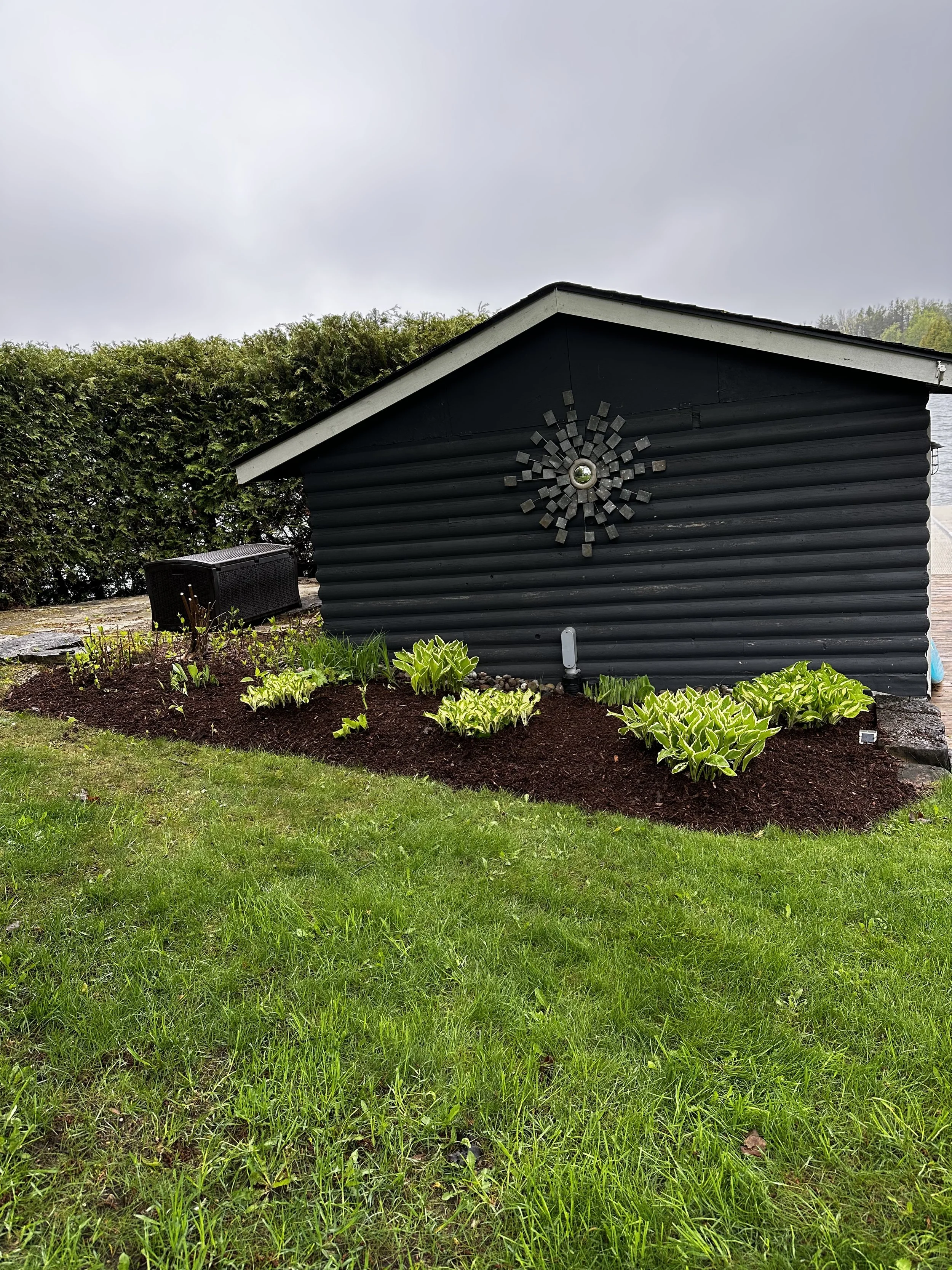 A black wooden shed with a decorative metal sunburst on its side, surrounded by green plants and grass, with a cloudy sky overhead.