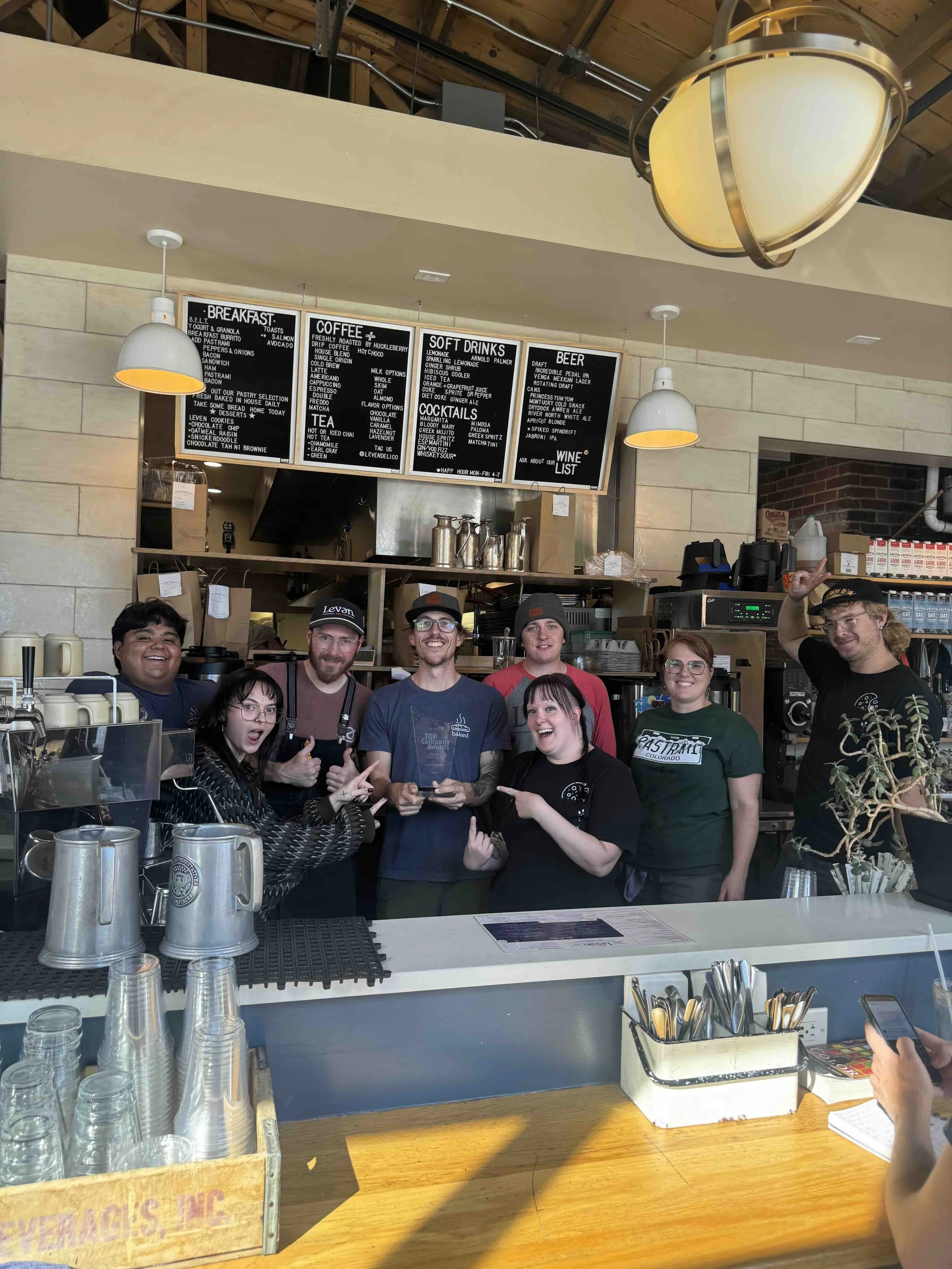 Group of staff members posing behind a counter in a cafe or restaurant, smiling and pointing at an award or plaque, with a menu board hanging above and barista equipment behind them.