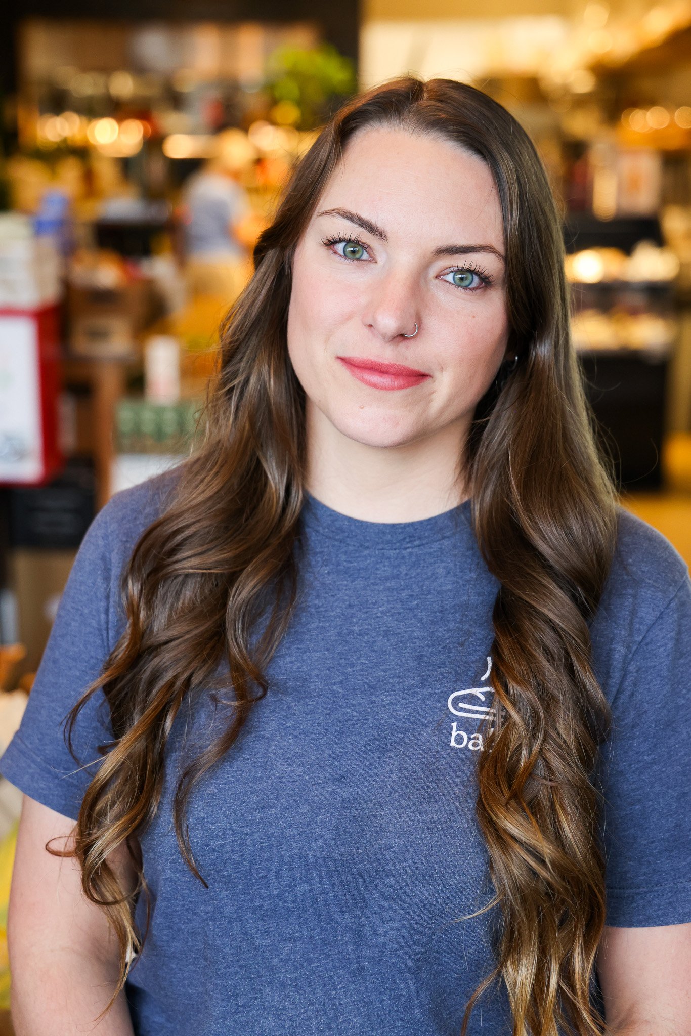 A young woman with long wavy brown hair, blue eyes, and a nose ring, smiling softly, wearing a blue t-shirt at Leven Supply