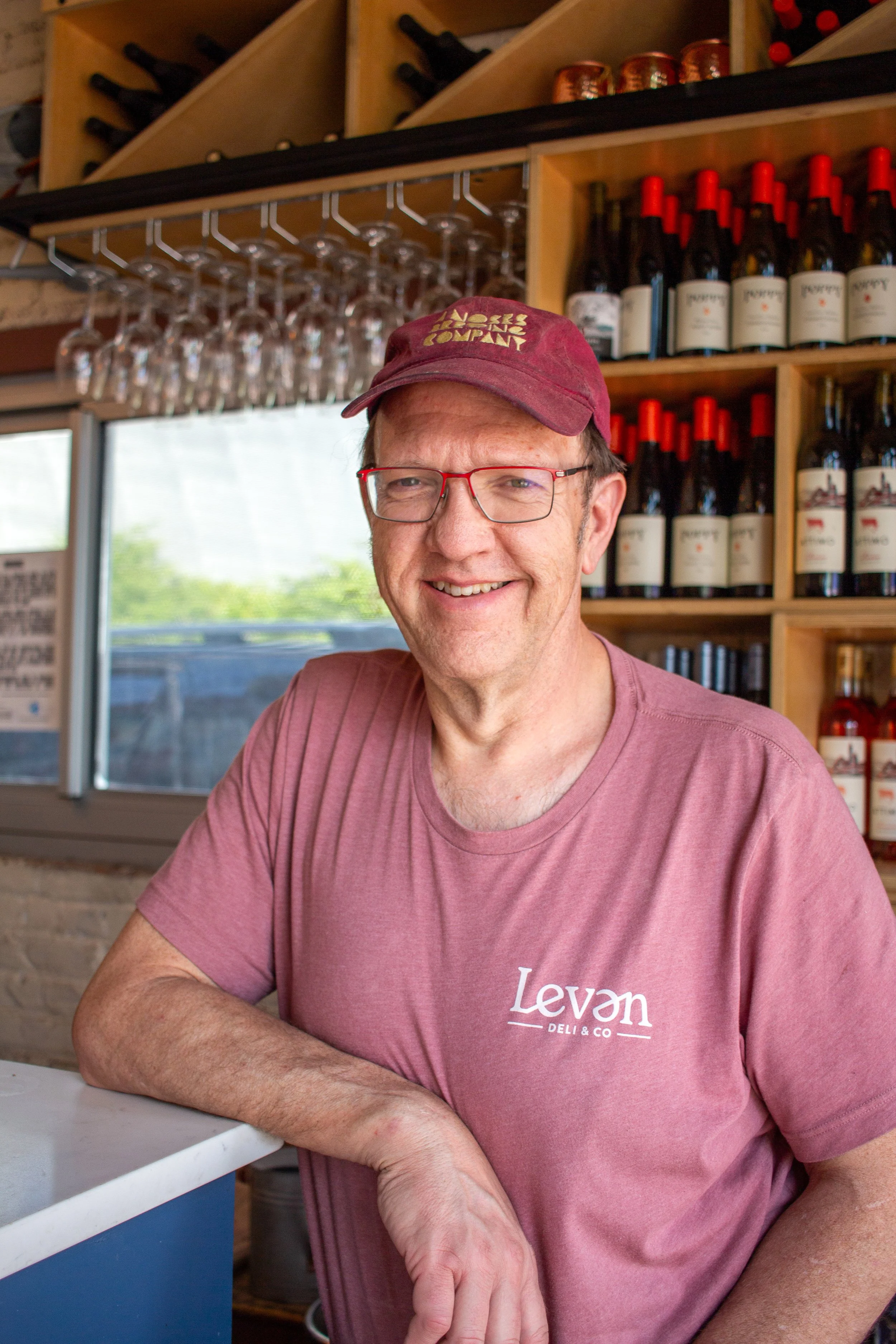 A smiling man with glasses and a maroon baseball cap standing inside a wine shop, with bottles of wine and wine glasses hanging upside down behind him.