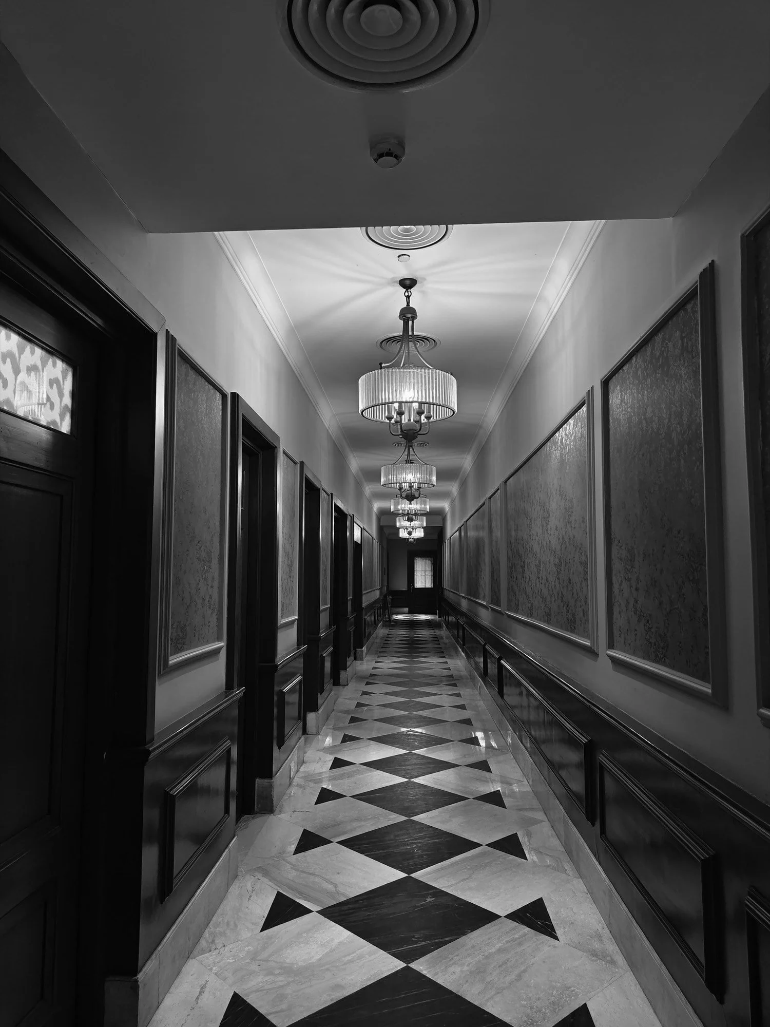 A long hallway of doors lit by hanging lamps, in black and white