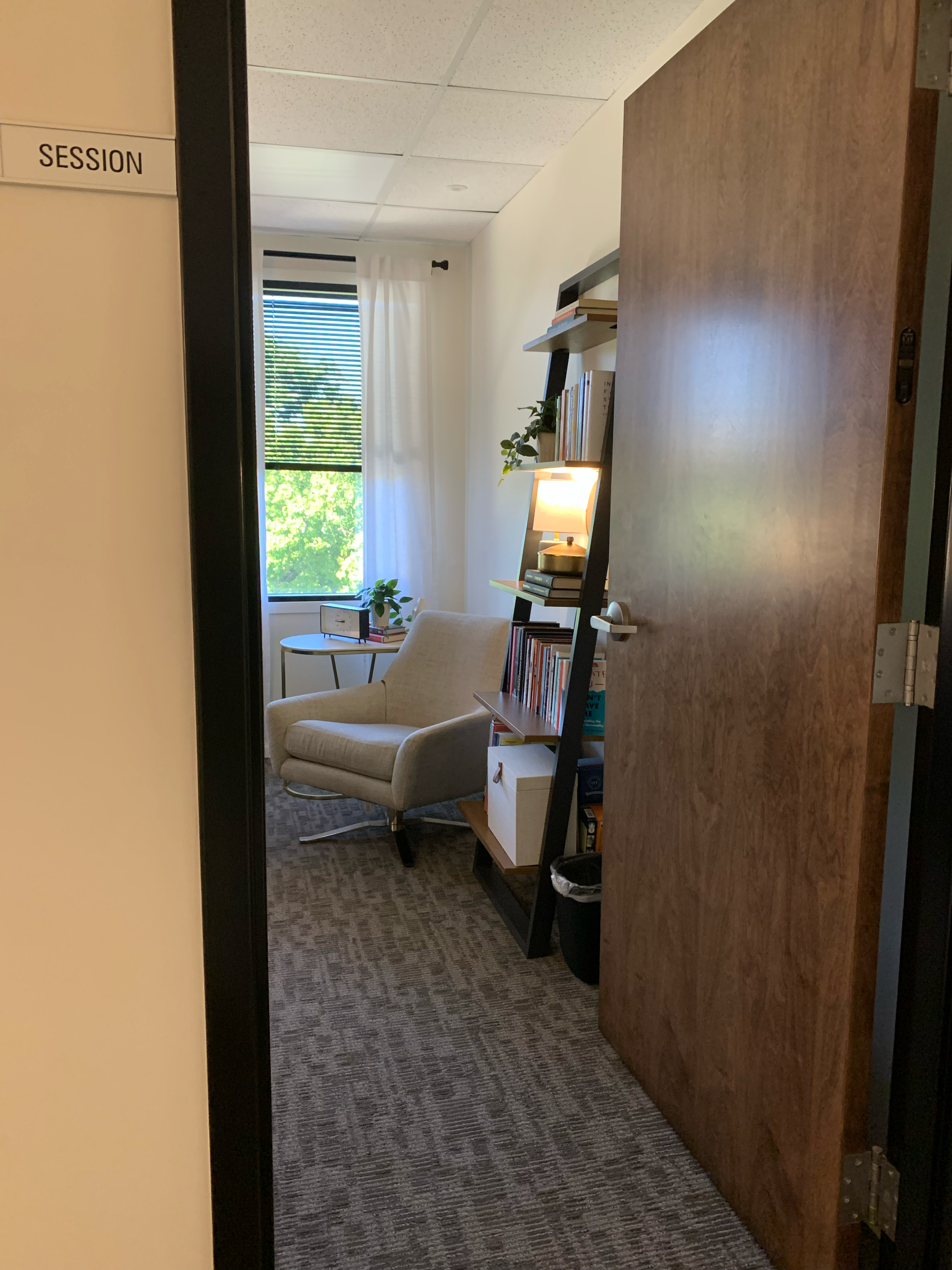 View of a cozy office space with a beige armchair near a window with blinds, a small round side table with a plant and laptop, a bookshelf with books and decorative items, and a lamp providing warm lighting.
