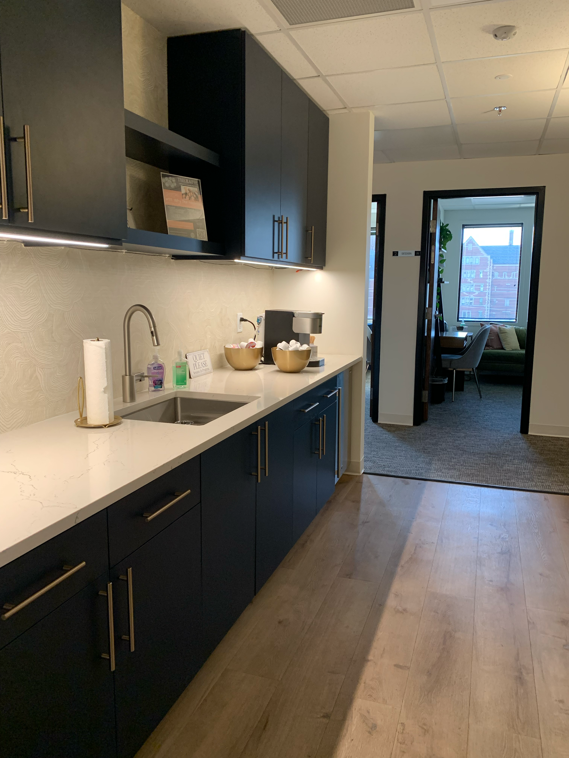 Modern kitchenette with navy blue cabinets and white marble countertop, kitchen sink, soap dispensers, and a coffee station. Adjacent room with a desk and sofa is visible through an open doorway.