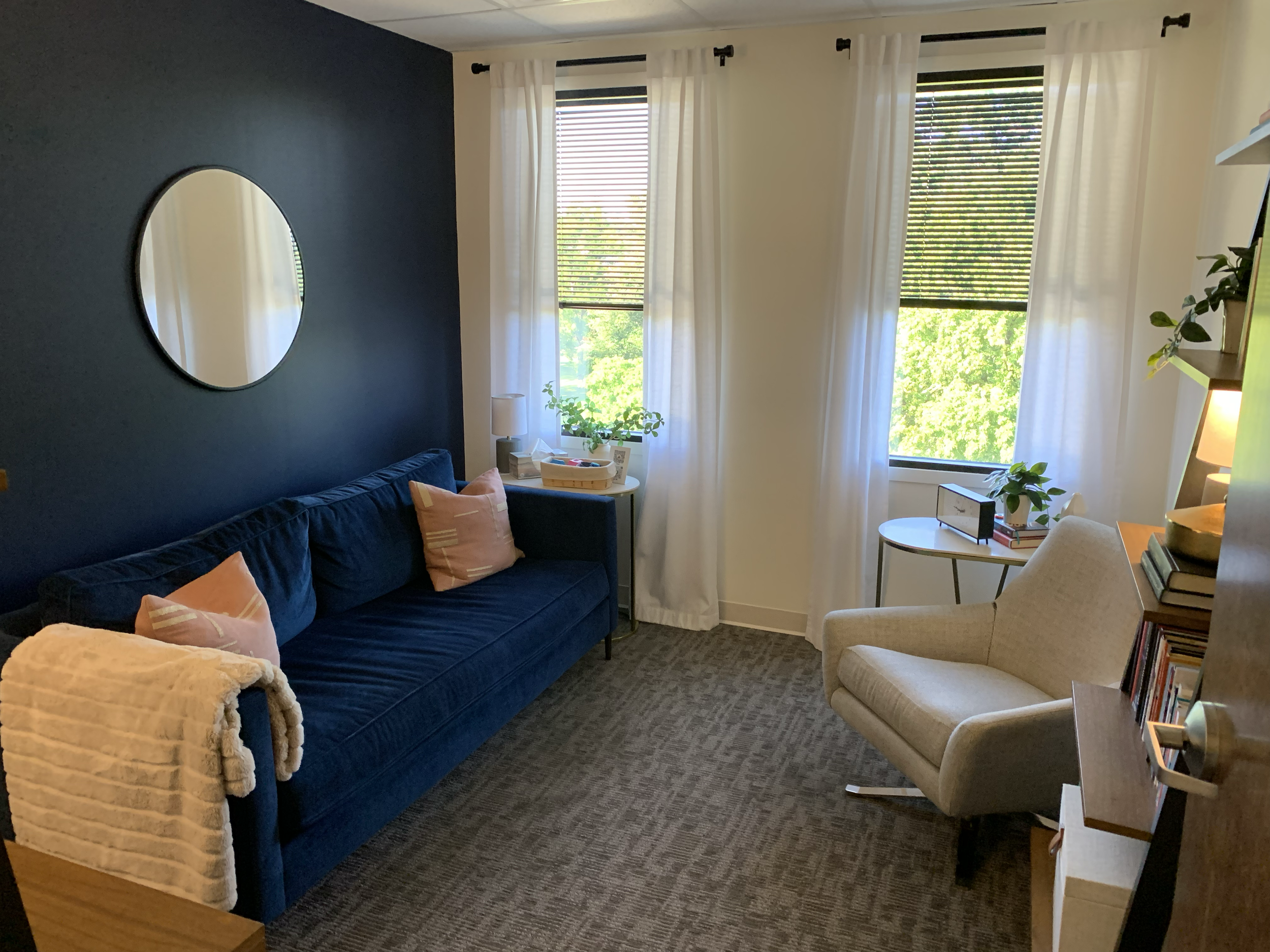A cozy living room with a navy blue couch, pink pillows, a round mirror on a blue accent wall, white curtains, and two windows with blinds showing green trees outside. There is a white armchair, a small side table with plants, books, and a lamp, and 