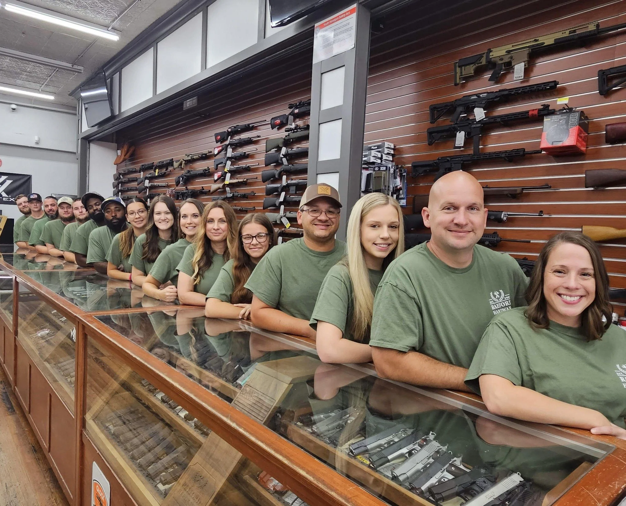 Group of staff standing in a row behind a glass display case filled with firearms in Raeford Hardware.