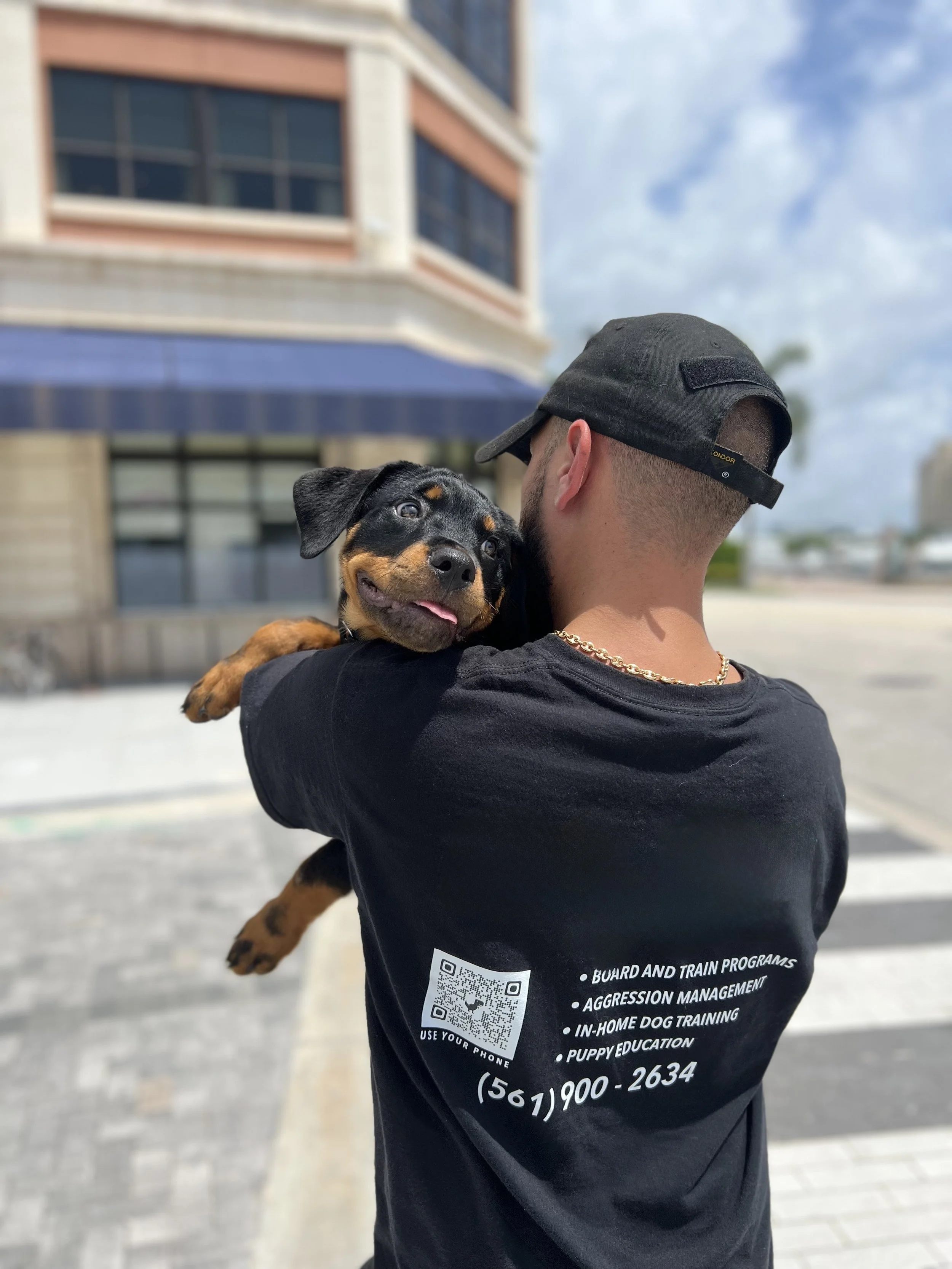 A man wearing a black cap and black T-shirt holding a smiling Rottweiler puppy with a cloudy sky and a building in the background.