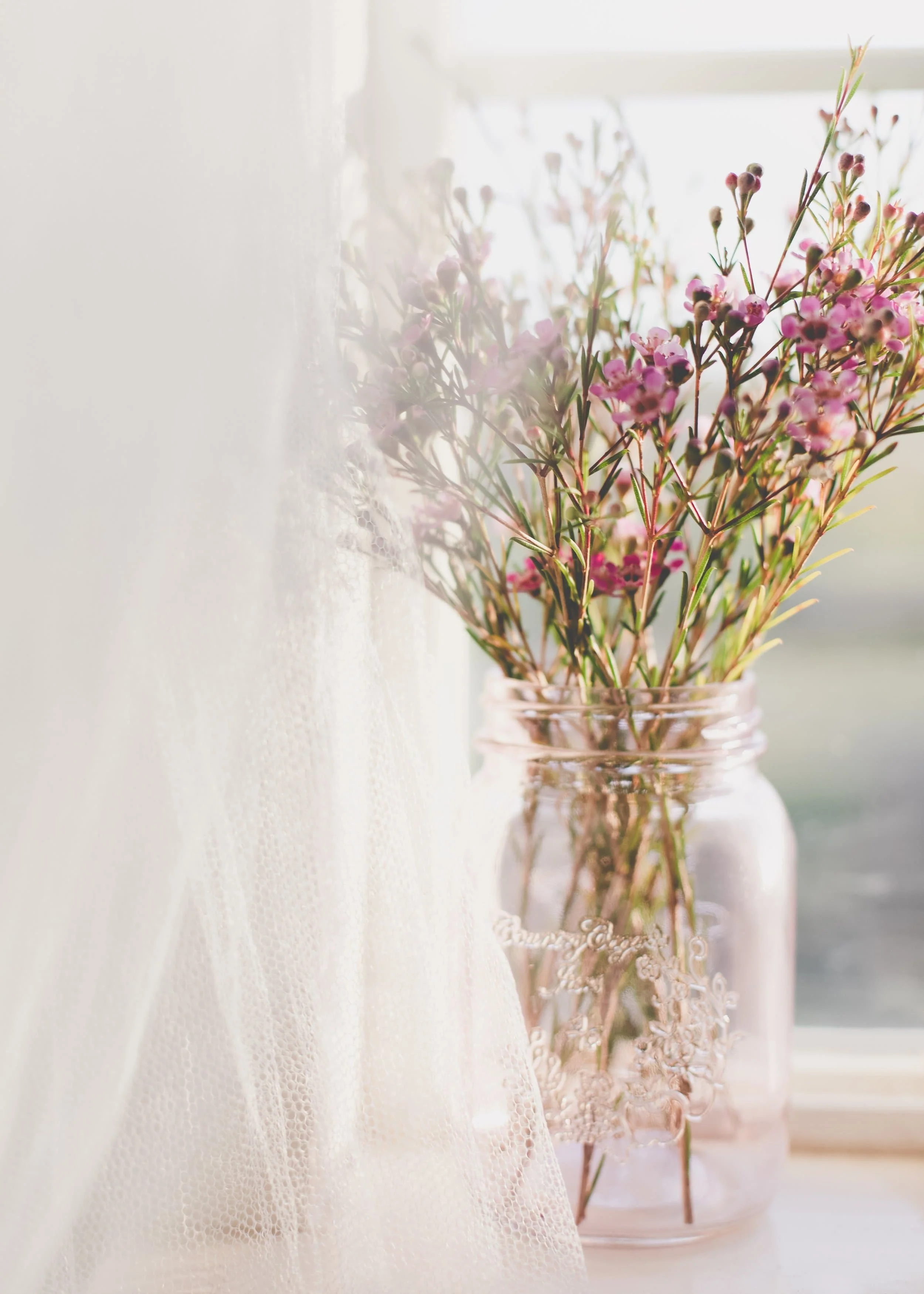 Pink wildflowers in a glass jar on a windowsill, partially obscured by a sheer curtain.