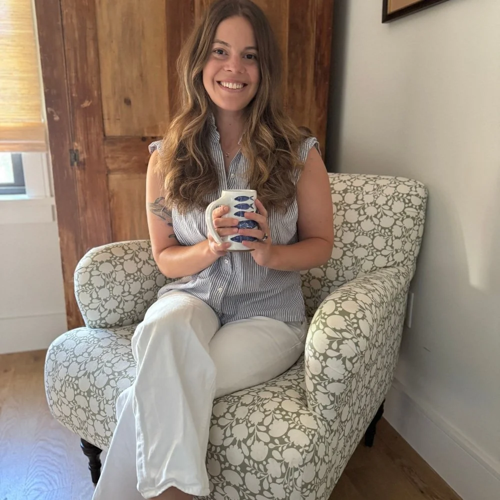 Abby Behar, Licensed Clinical Social Worker, specializing in trauma and anxiety treatment, woman with long wavy brown hair and a striped sleeveless top sitting on a floral patterned armchair, holding a mug with fish designs, smiling at the camera.