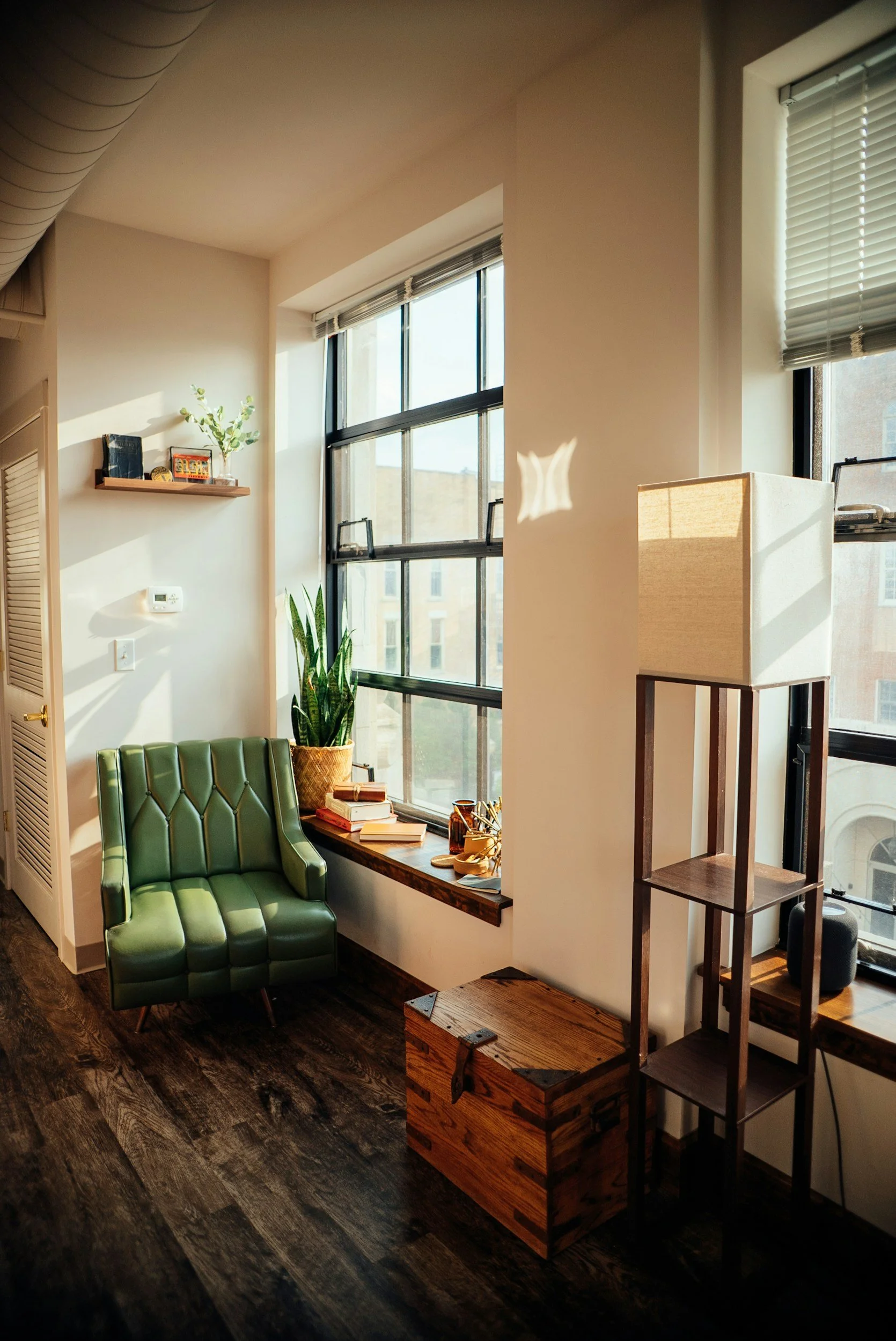 A small, sunny corner of an apartment with a green armchair by the window, a wooden chest, and open shelves