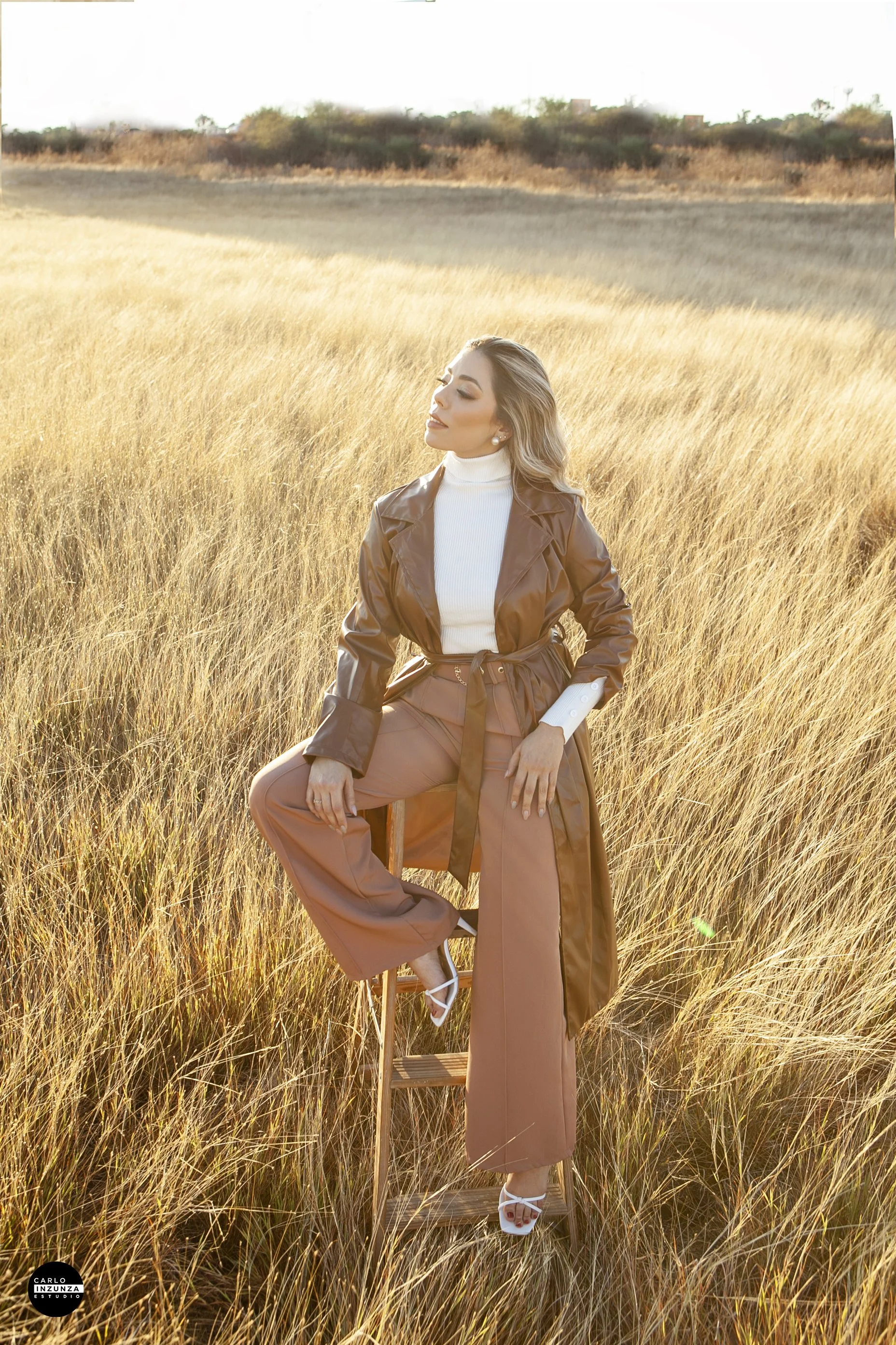 A woman sitting on a wooden stool in a field of tall, golden grass during sunset. She is wearing a tan trench coat, white turtleneck, wide-leg pants, and white heels, with her eyes closed and a peaceful expression.