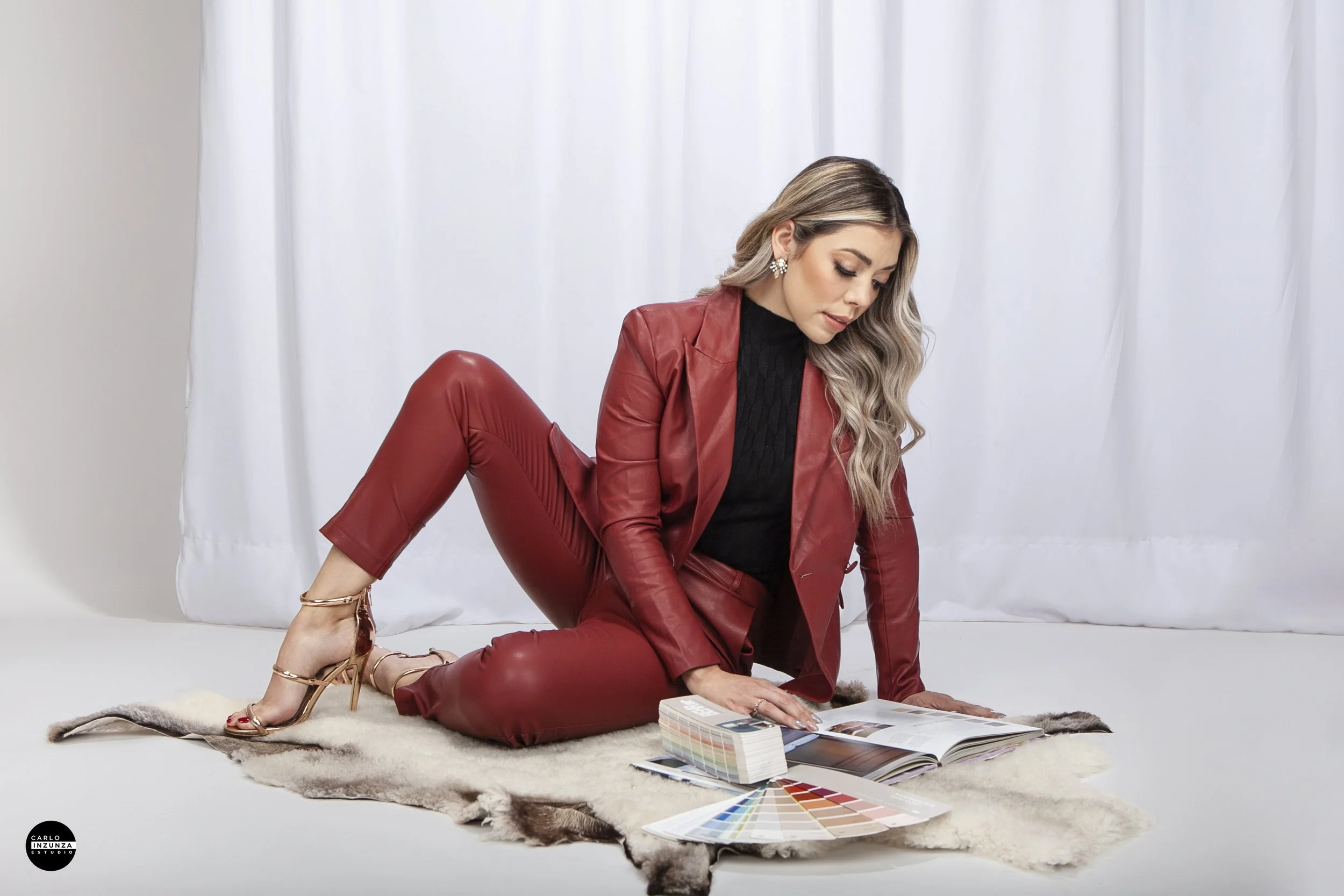 Woman in red leather suit sits on a fur rug looking at fabric color samples and a design catalog in a studio with white curtains.