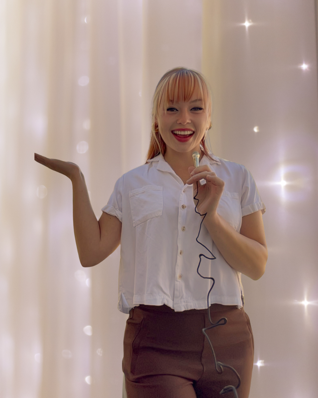 A dedicated and certified life coach near me holding a microphone and smiling, standing in front of a light-colored curtain with small light spots, providing advice on self-talk, inner critics, and career advancement as an empowerment coach.