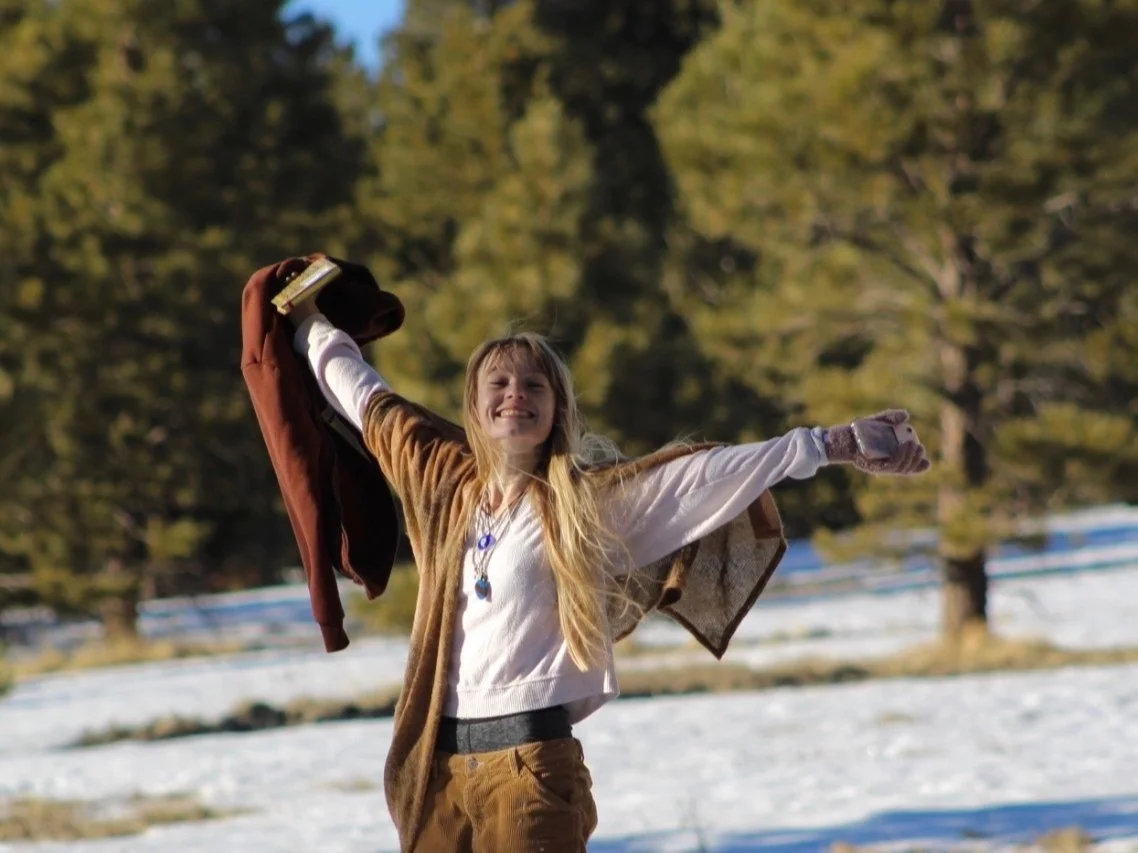 Young girl with long blonde hair smiling and warmly stretching her arms out outdoors on a bright, sunny day, with a self care journal in hand, pine trees and snow-covered ground of the Coconino national forest in Flagstaff Arizona in the background.