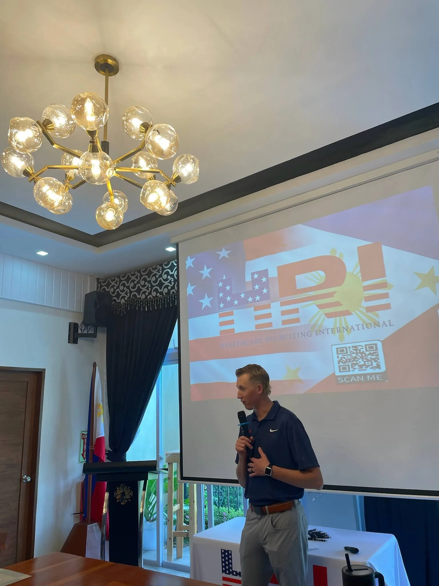 Dawson Mangum giving a presentation at an international healthcare event, standing near a large screen displaying the logo HRI (Healthcare Recruiting International) with American and Filipino flags, and a QR code. The room has a Philippine flag.