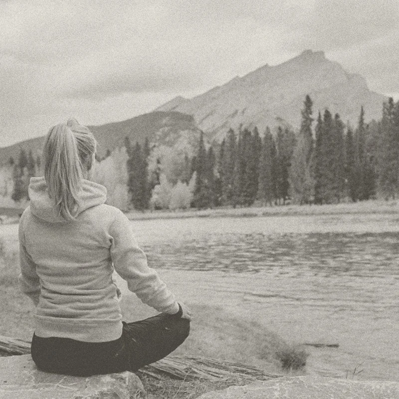 A woman sitting cross-legged by a body of water, facing away, with mountains and trees in the background.