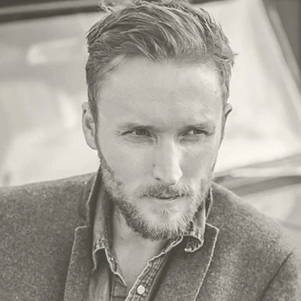 A black and white photo of a man with styled hair, beard, and mustache, wearing a collared shirt and blazer, looking to the side.