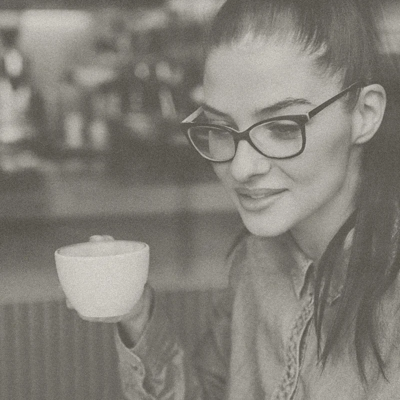 A woman with glasses holding a coffee cup, smiling in a cozy cafe setting.
