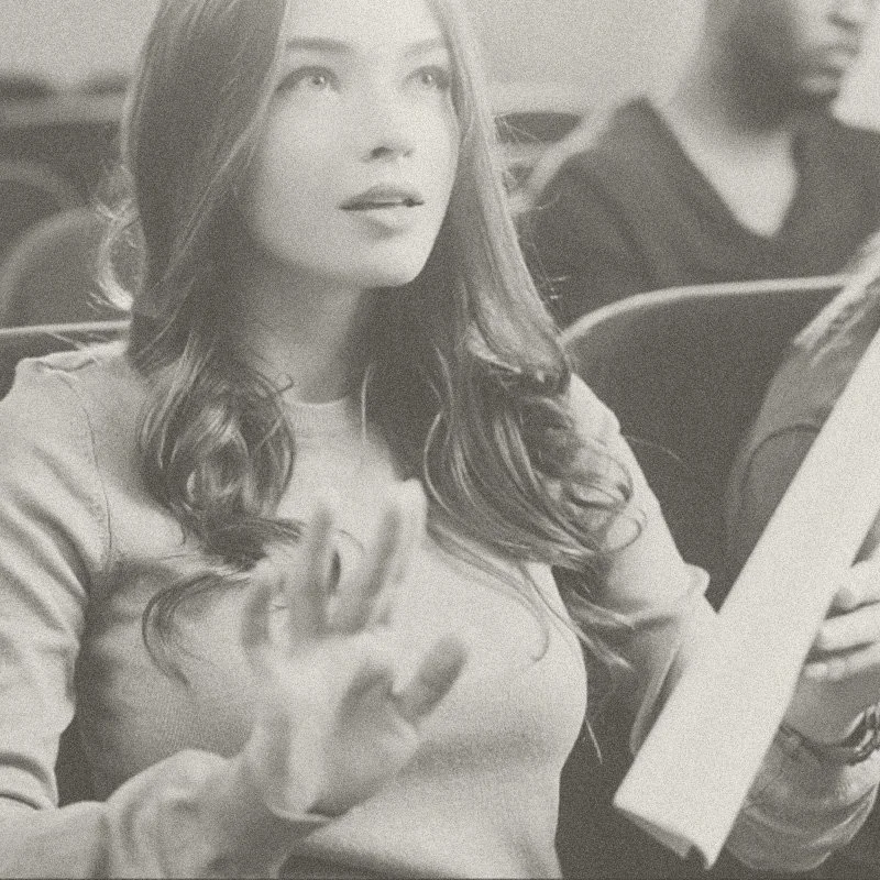 A woman with long hair is sitting while holding a book, with other people visible in the background.