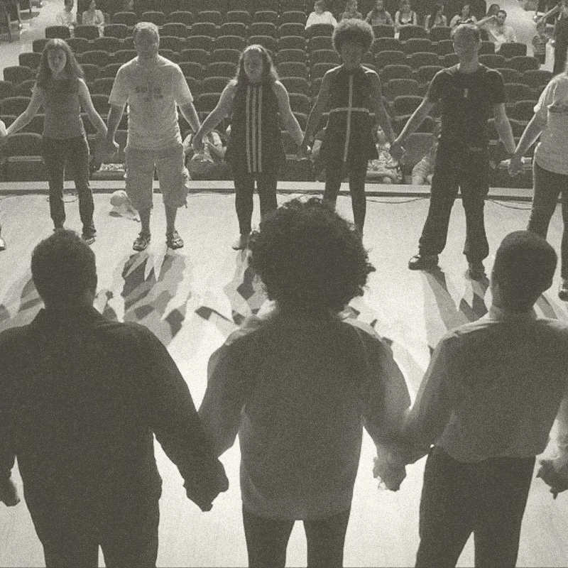 People holding hands in a circle on a stage, facing an audience.
