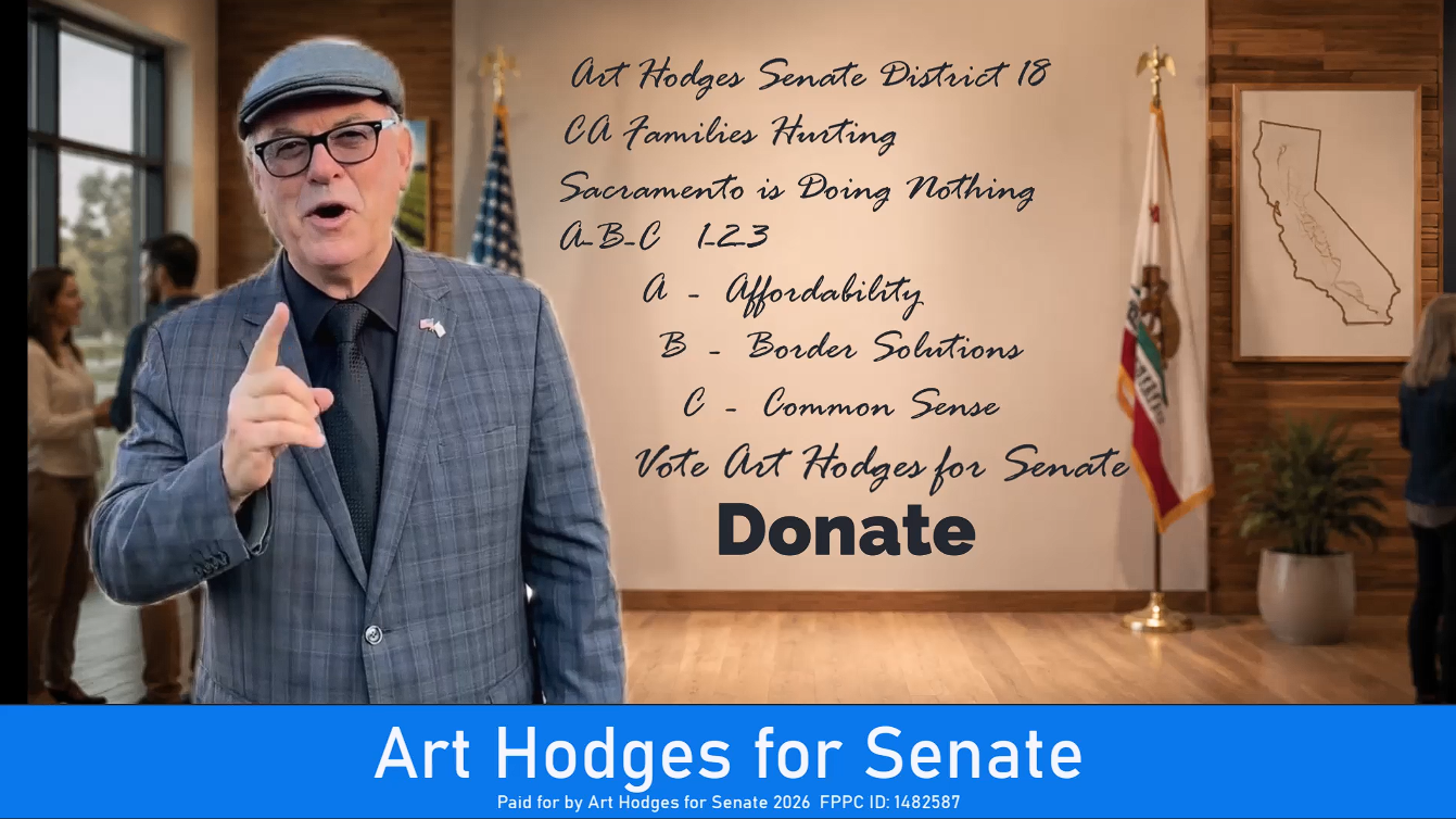 A man in a gray plaid suit, glasses, and a hat standing in a campaign office, pointing upward. Behind him, a wall with a large handwritten-style sign promoting Art Hodges for Senate, listing his district, positions, and a call to donate. The office has large windows, with people in the background, and flags on the right side.