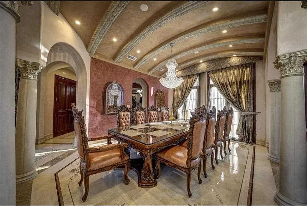 Elegant dining room with a large wooden table, ornate upholstered chairs, a chandelier, tall windows with heavy drapes, and decorative columns.
