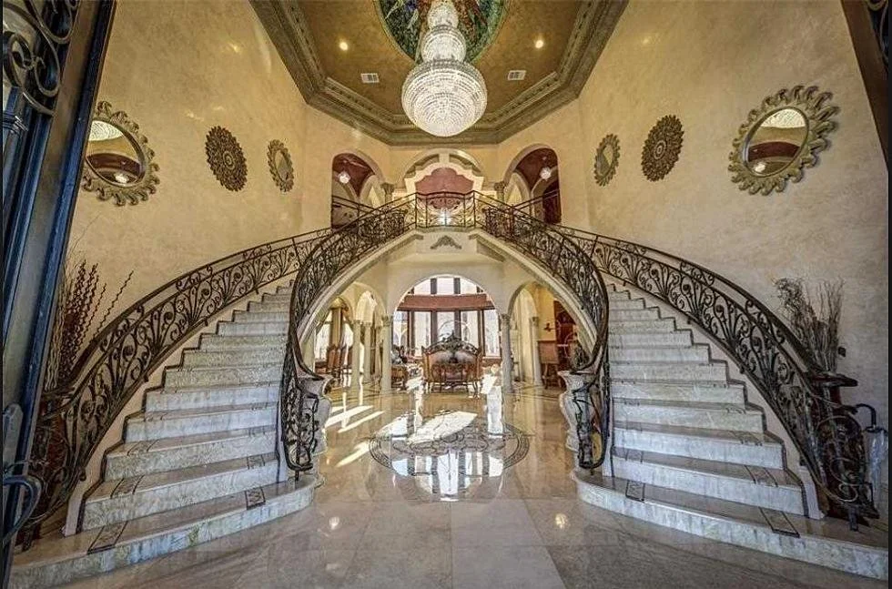 A grand foyer with a marble staircase curving upward on both sides, ornate wrought iron railings, a large chandelier hanging from a decorative ceiling, and walls adorned with round mirrors and decorative round wall hangings.